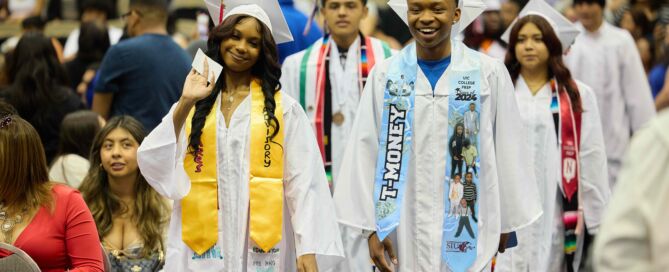 UIC College Prep graduates walk down the aisle in white graduation robes. They are smiling.