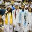 UIC College Prep graduates walk down the aisle in white graduation robes. They are smiling.