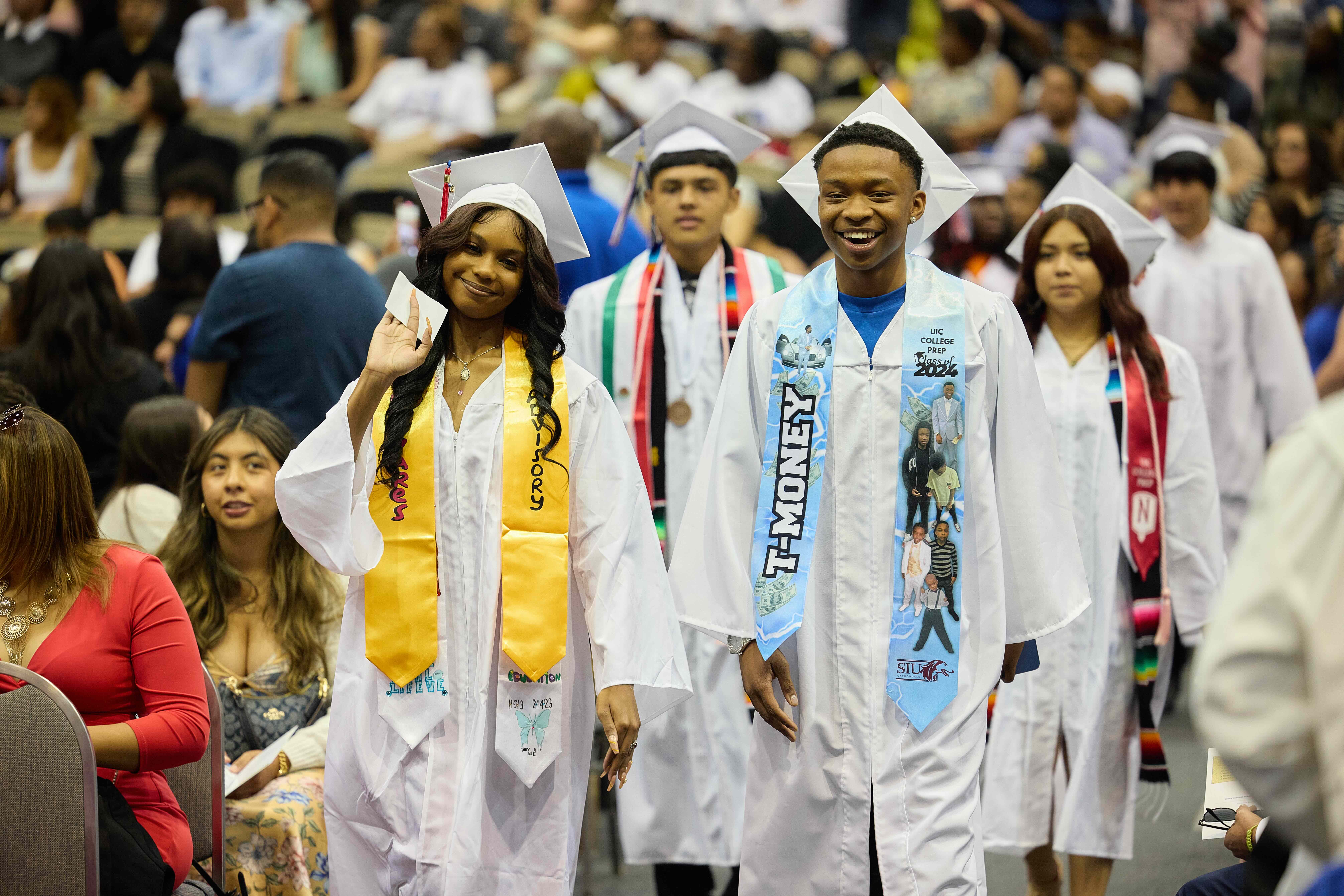 UIC College Prep graduates walk down the aisle in white graduation robes. They are smiling.