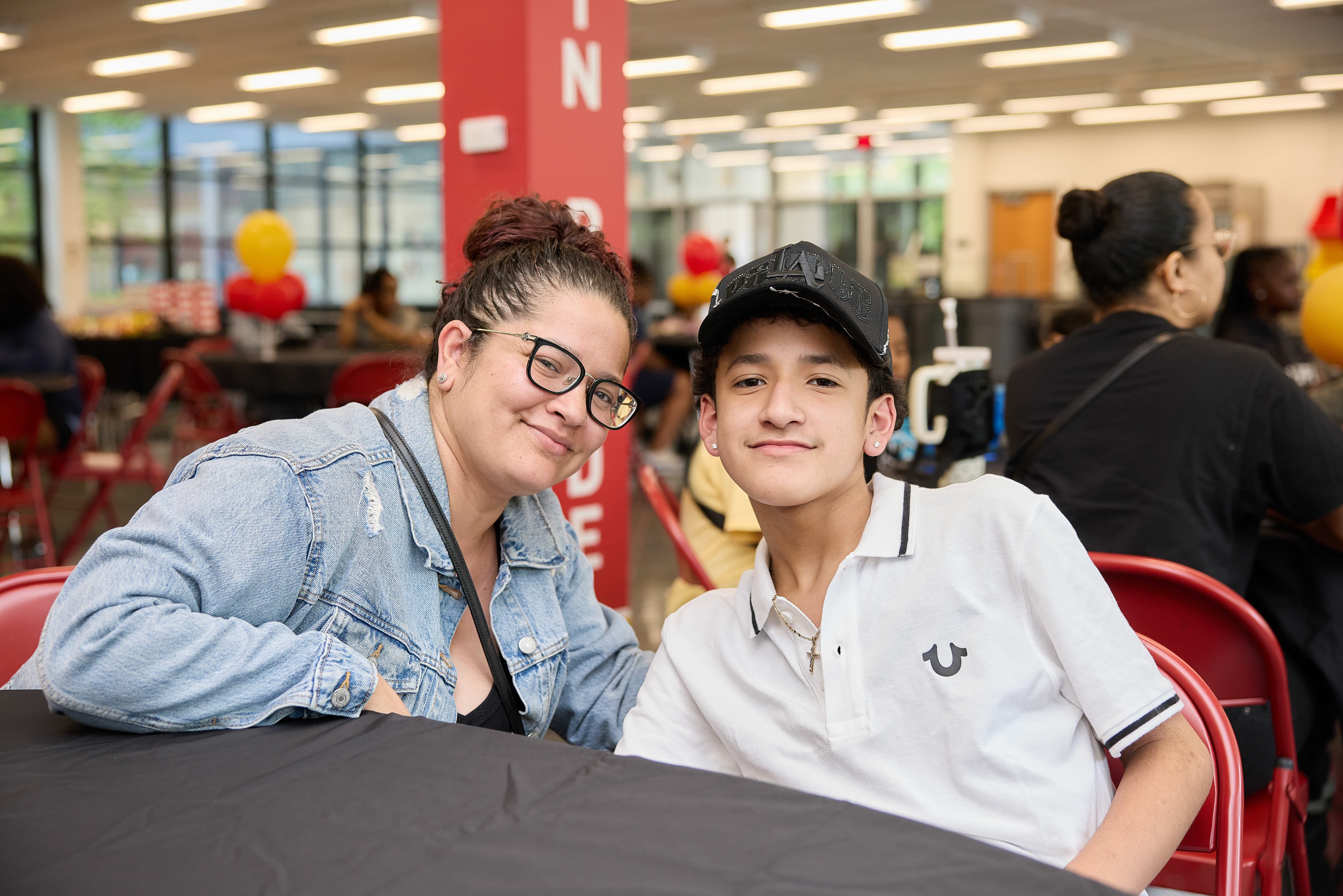A parent and their kid pose for a photo at Freshman Signing Night in The Noble Academy's cafeteria. They are smiling and you can see a crowd of other families and kids behind them.