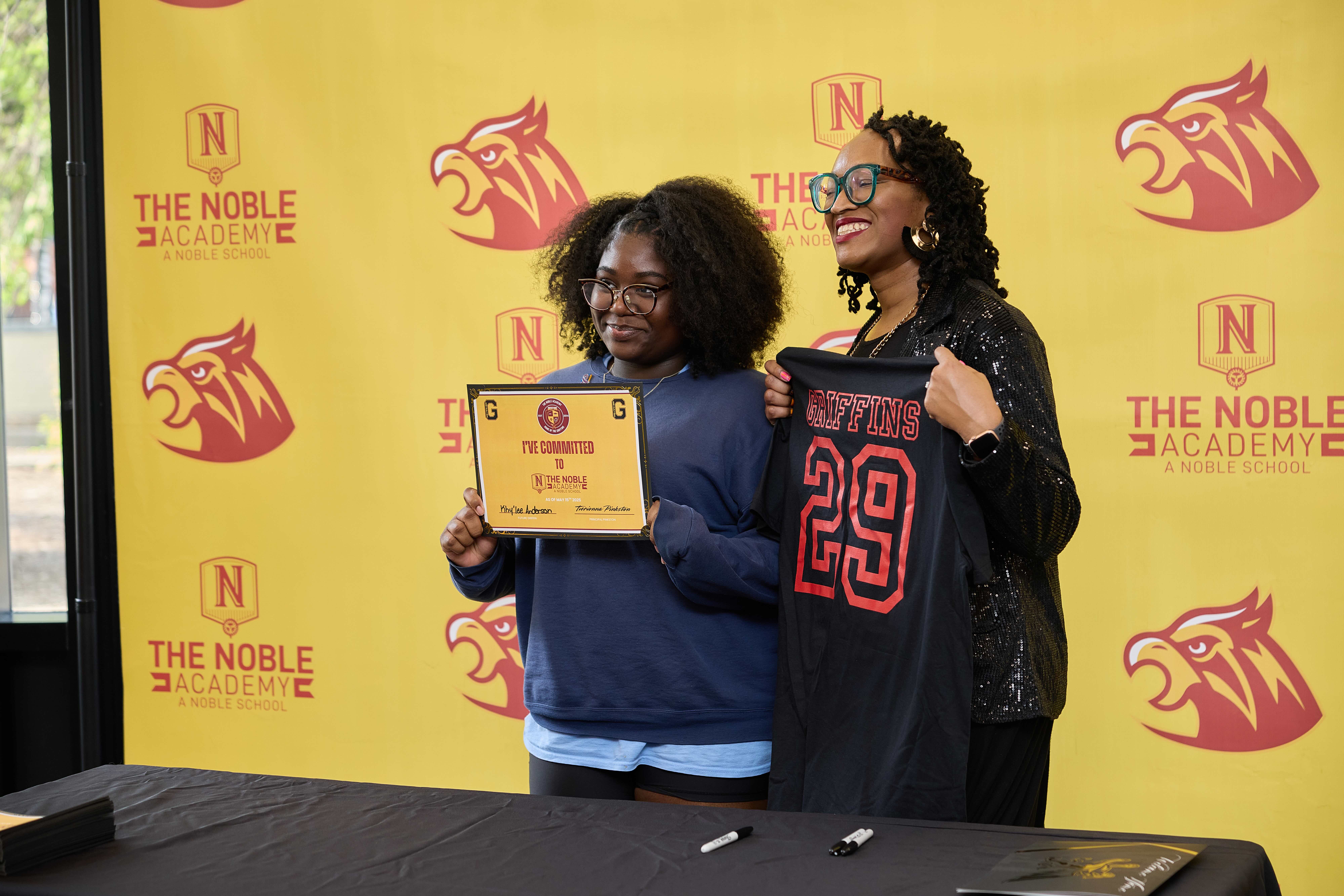 An incoming TNA freshman poses for a photo with Principal Pinkston with her "I'm Committed to TNA" certificate in hand. You can see a yellow and red photo backdrop behind them with TNA's griffin mascot on it.