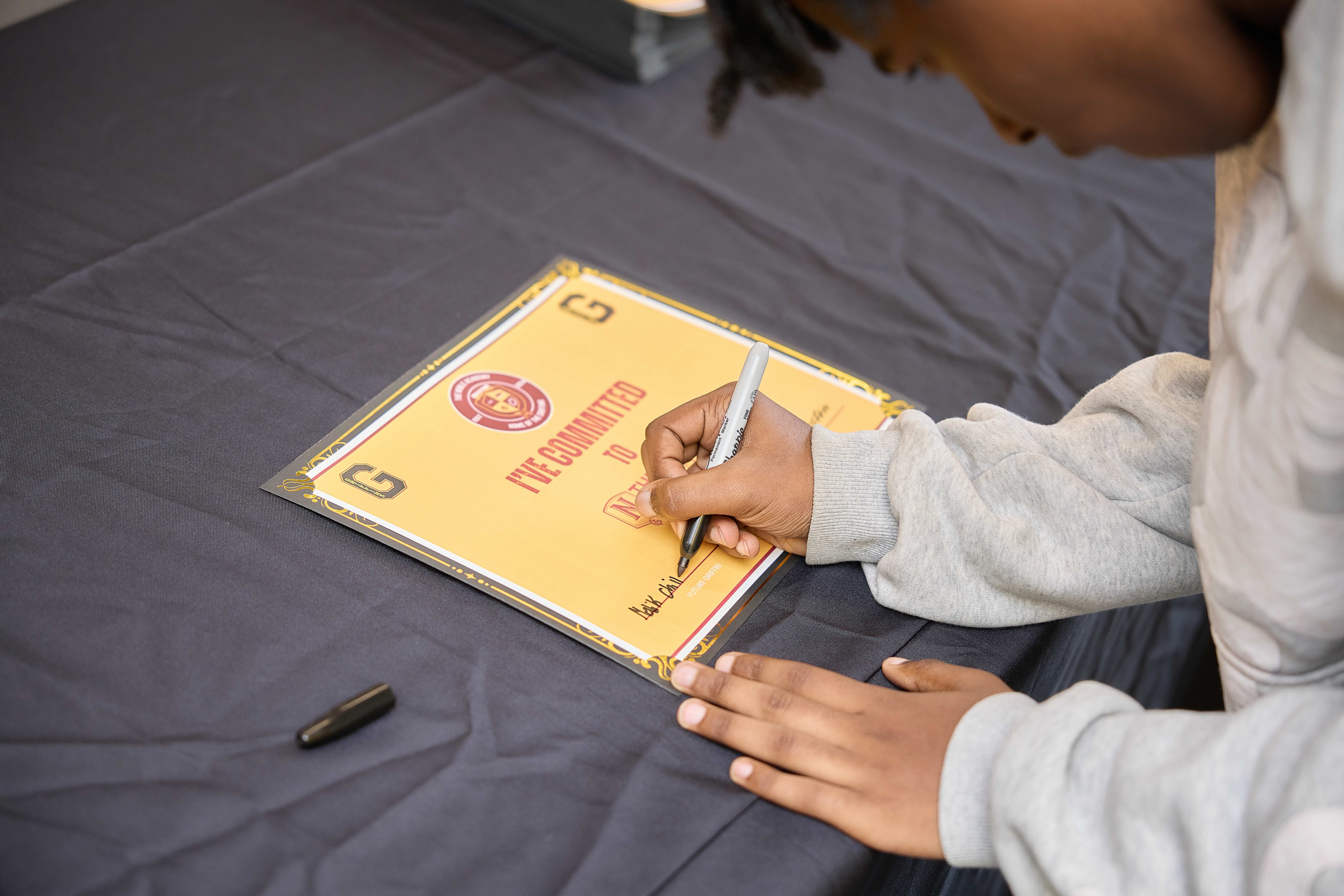 An incoming freshman signs a yellow and red certificate that says "I'm committed to..." followed by The Noble Academy logo.