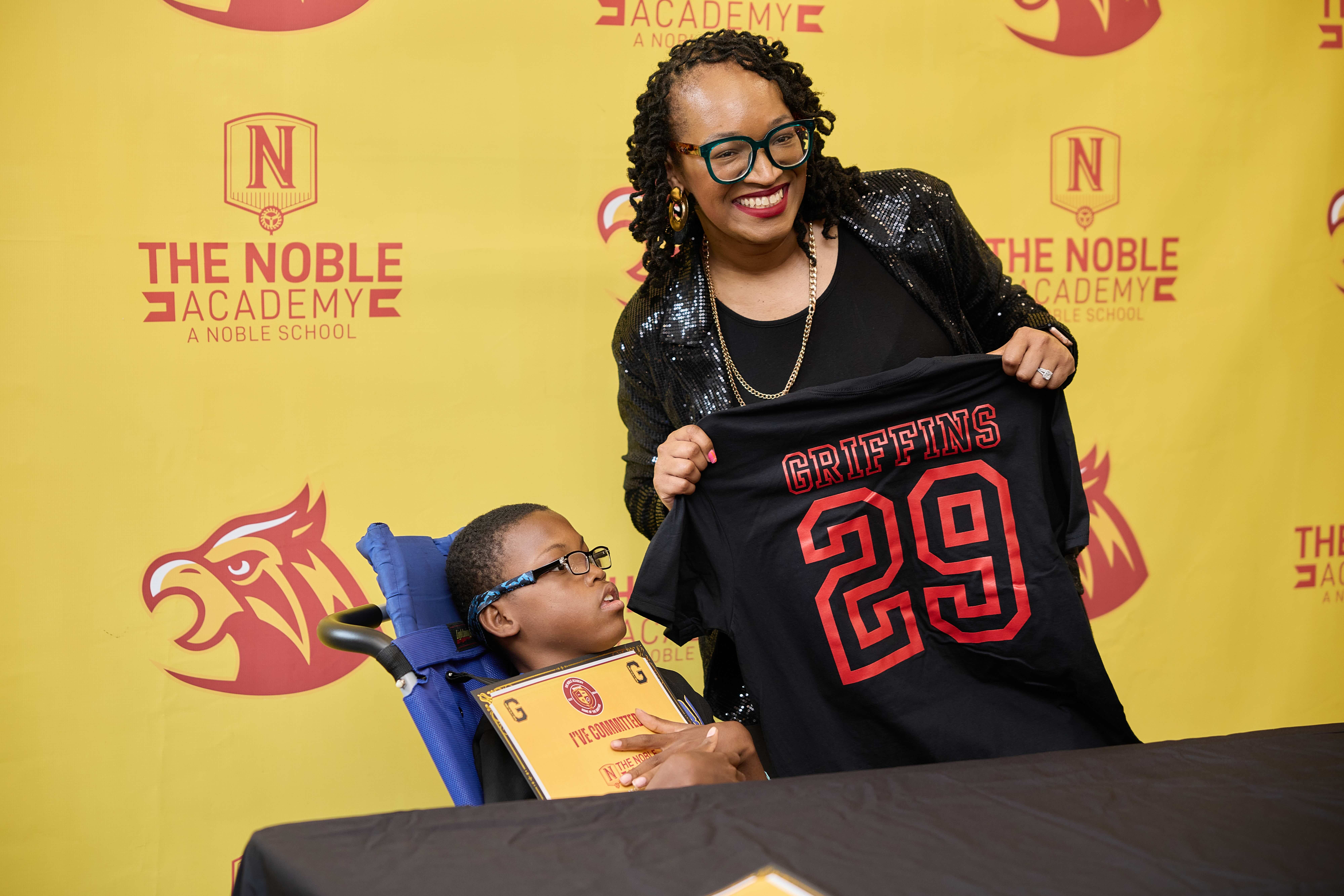 An incoming freshman in a wheelchair holds their "I'm Committed" certificate while posing for a photo with Principal Pinkston. You can see the yellow and red photo backdrop behind them.
