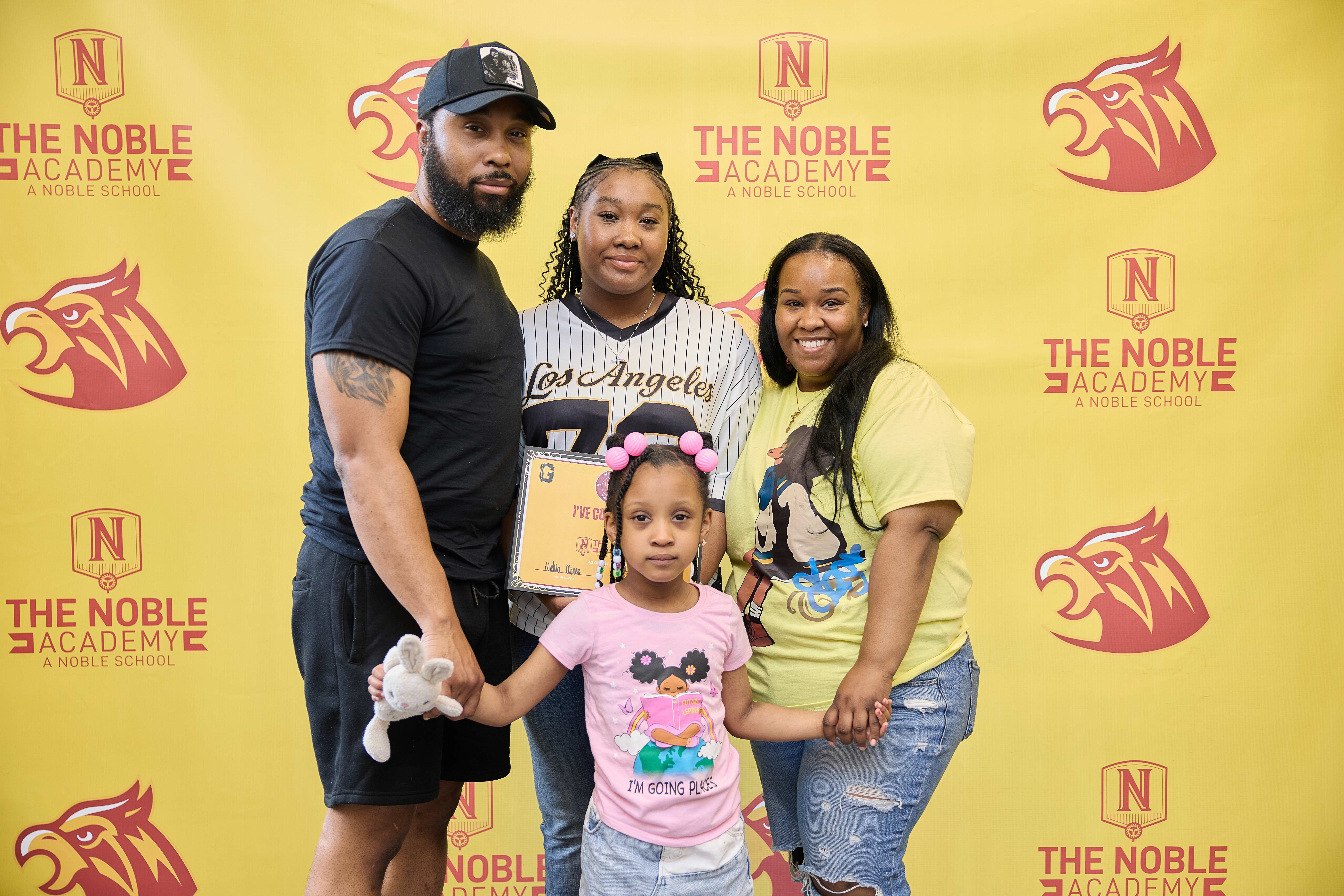 An incoming TNA freshman with their family stands in front of the yellow and red photo backdrop. You can see the student, their parents, and their younger sibling. The student holds their certificate.
