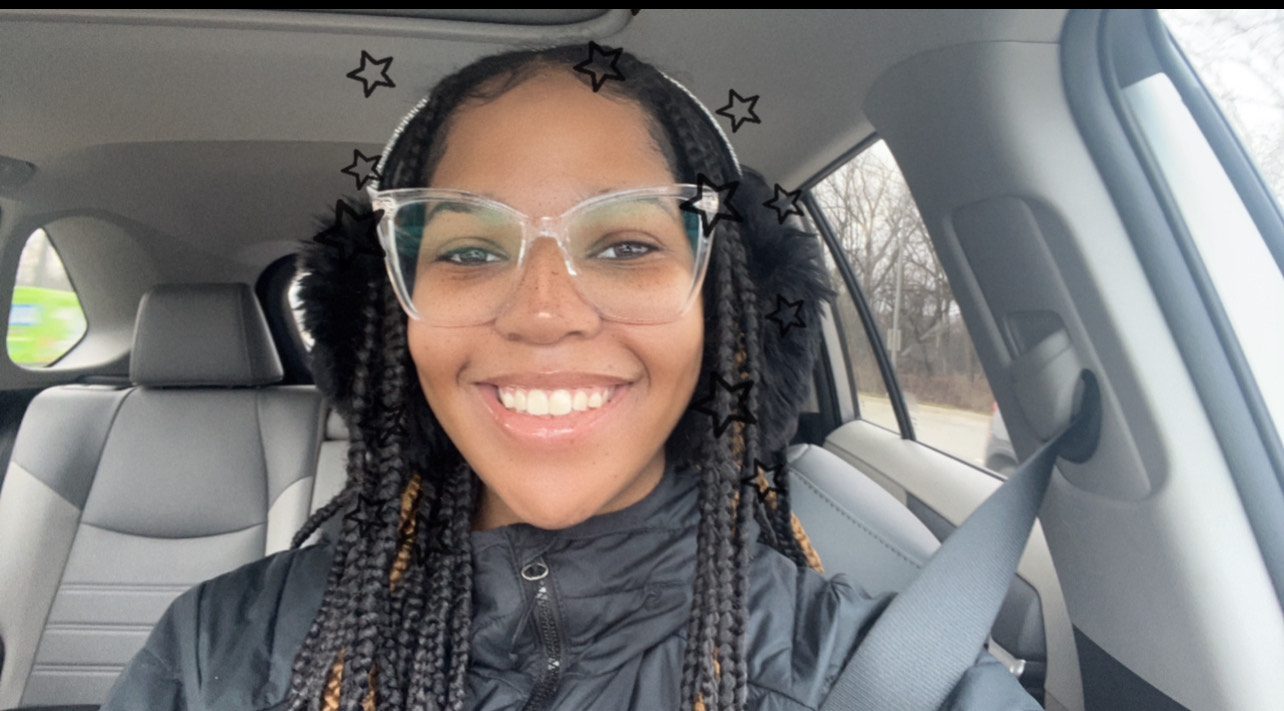 Amira Amira Todd sits in a car, taking a selfie. She is a young woman with long black braids, freckles, and light brown skin. She is smiling and wearing glasses, fuzzy black ear muffs, and a black coat.