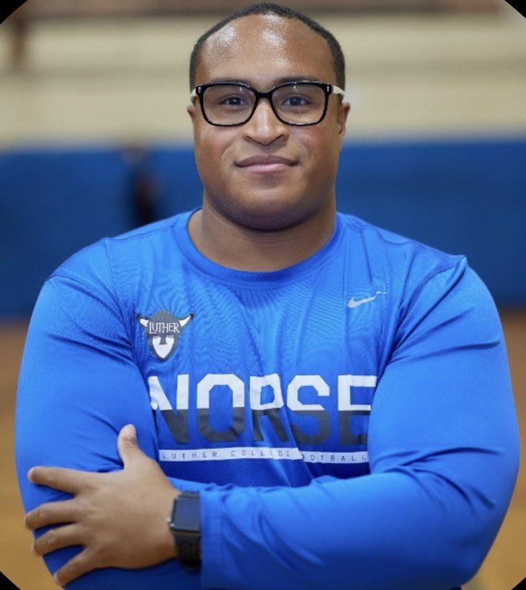 CoachDiaz (1) A headshot of Coach Diaz. He is a man with short buzzed black hair and medium-dark brown skin. He is crossing his arms and wearing glasses and a light blue athletic long-sleeved shirt with the Luther College football logo and mascot on it.