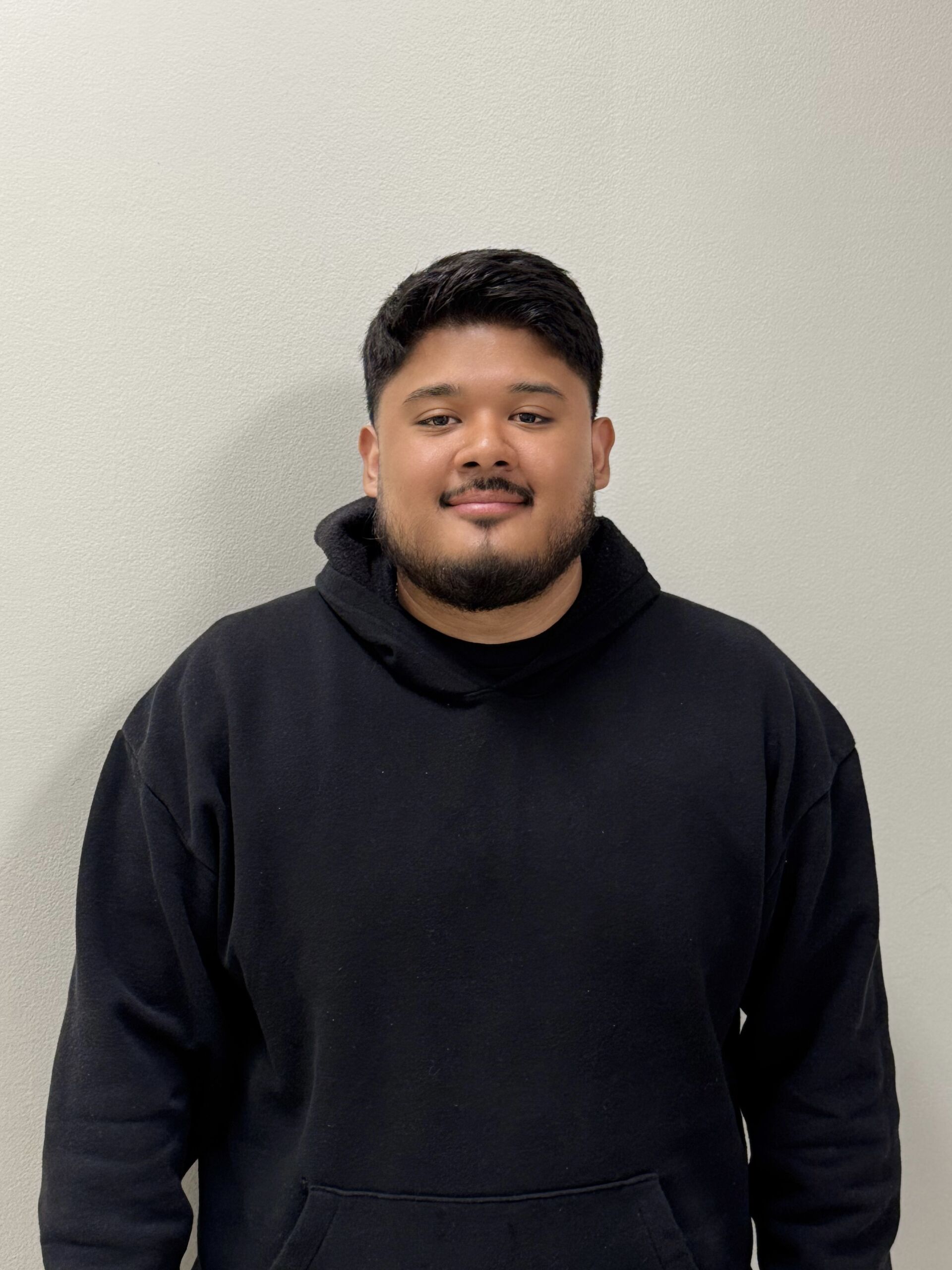 CoachRamirez (1) A headshot of Edwin Ramirez. He is a man with short black hair, a full beard, and light brown skin. He is smiling and wearing a black hoodie and standing in front of a white wall.