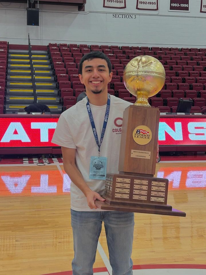 Luis stands in a gym, holding a massive trophy with a golden basketball on top. The trophy says "Patriot League Champions" and has all the names of Luis' team under that. Luis is a young man with short dark brown hair and brown skin. He is smiling brightly and wearing a gray Colgate University t-shirt with jeans.