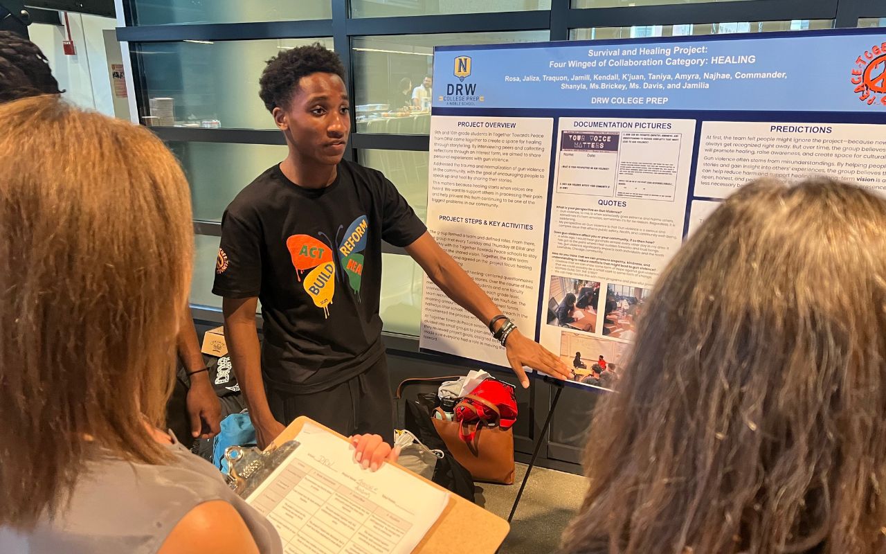DRW TTP 2025 Blog Cover Jamill Willis, a student at DRW College Prep, points to a posterboard with his group's Together Towards Peace project, which addresses gun violence in our communities.