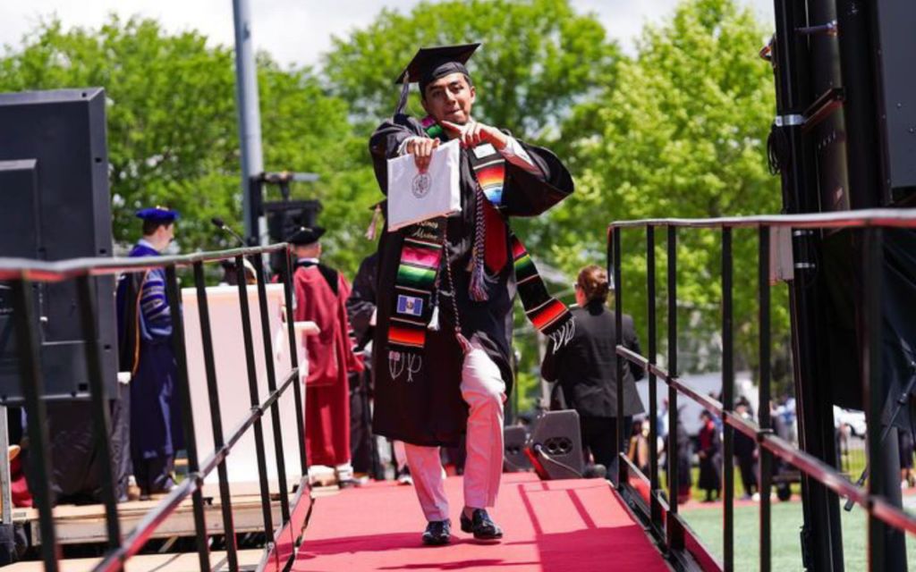 Luis Ramos points confidently at the camera, college diploma in hand and decked out in his graduation robes, as he walks off the stage. He is a young man with short dark brown hair, a light beard, and brown skin.
