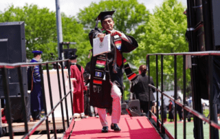 Luis Ramos points confidently at the camera, college diploma in hand and decked out in his graduation robes, as he walks off the stage. He is a young man with short dark brown hair, a light beard, and brown skin.