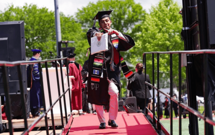 Luis Ramos points confidently at the camera, college diploma in hand and decked out in his graduation robes, as he walks off the stage. He is a young man with short dark brown hair, a light beard, and brown skin.