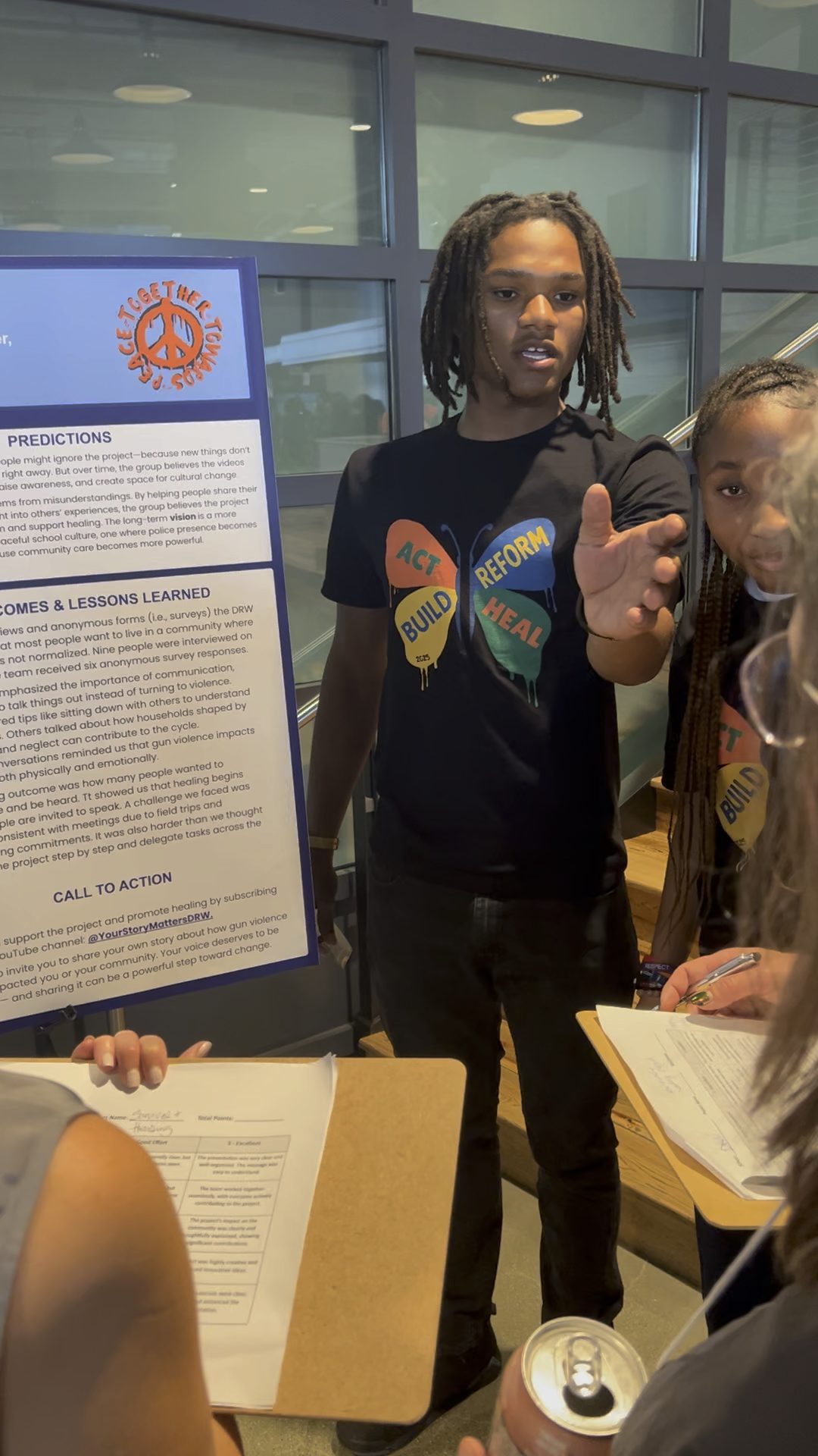 Kjuan Newby presenting Kjuan holds his hand up to address a audience member as he presents his group's project on a posterboard behind him. He is a young man with medium-length dark brown locs and dark brown skin. He is wearing one of the butterfly t-shirts.