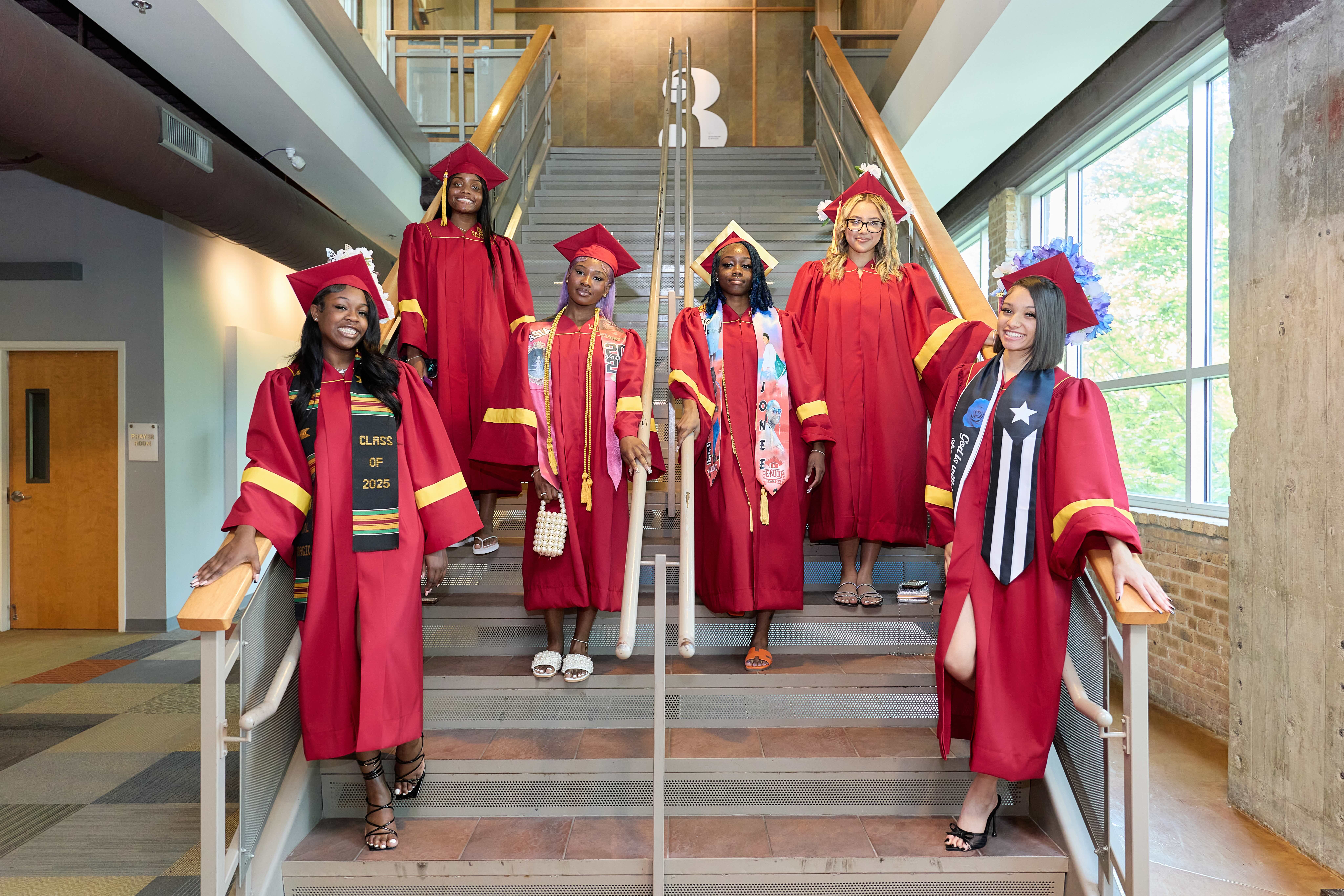 A group of six seniors pose on the stairs after their graduation. They are all decked out in bright red graduation robes with personalized sashes.