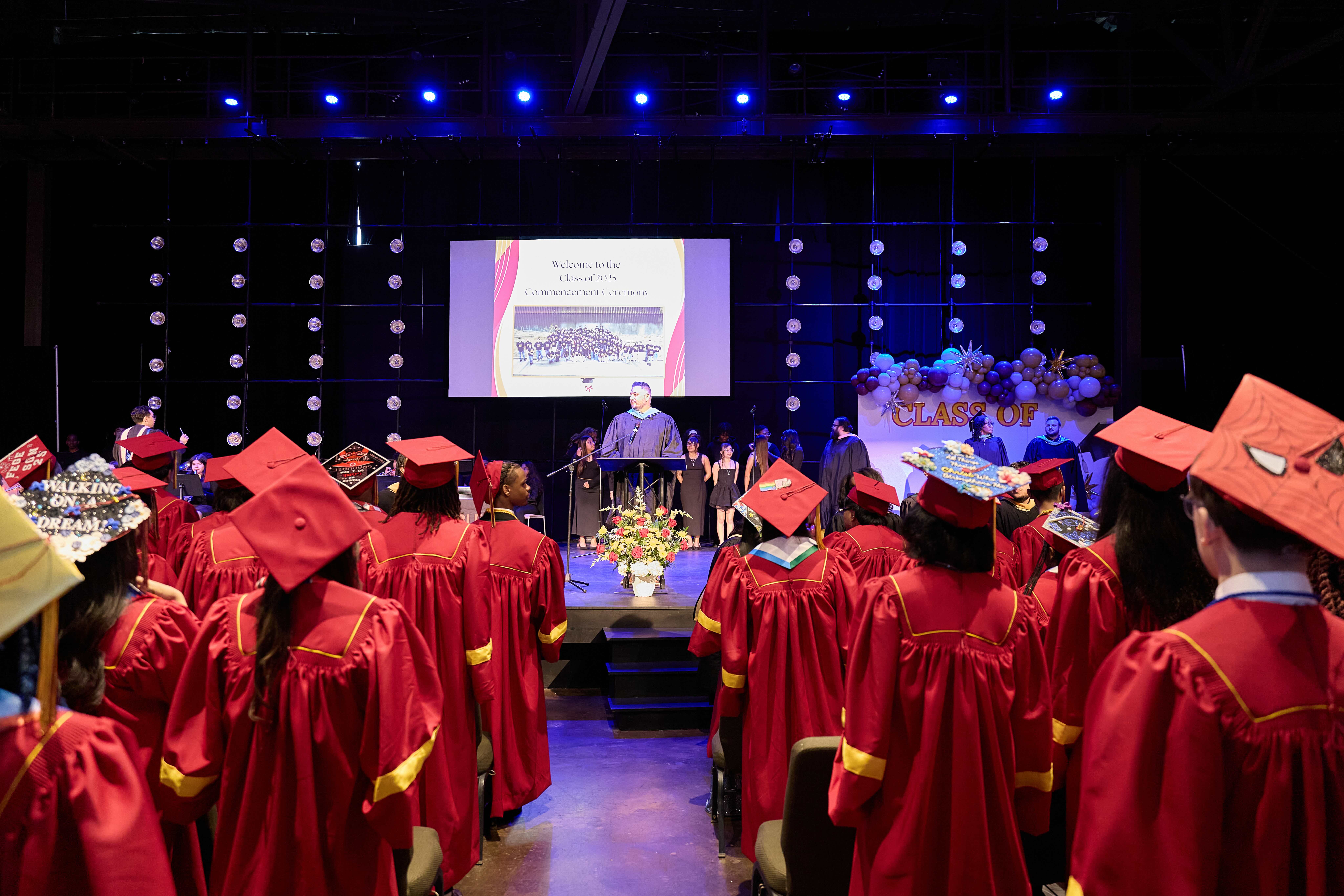 The Noble Academy's Class of 2025 stands up in their graduation robes while they are welcomed by a staff member on the stage.