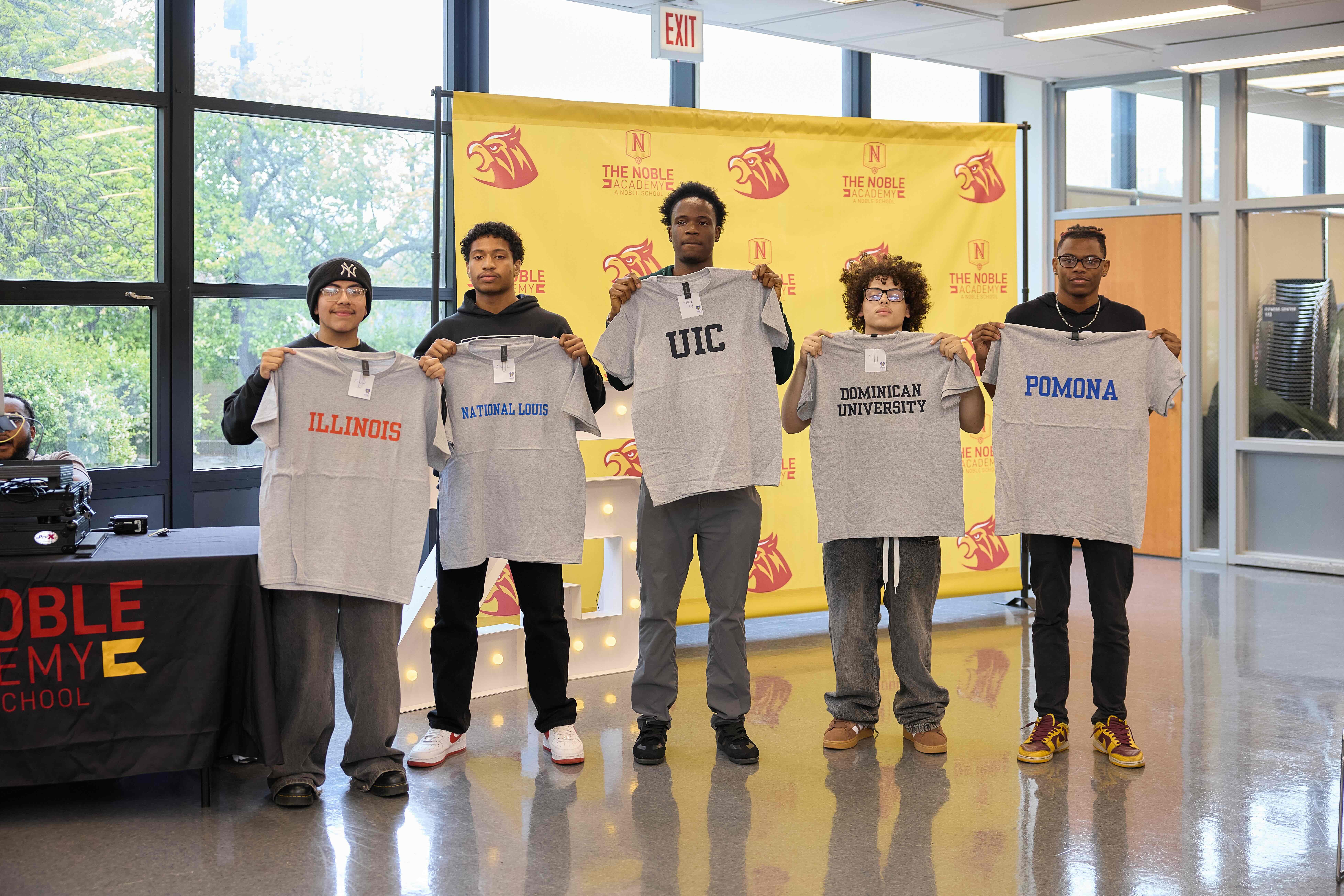 A group of 5 TNA seniors stand with gray shirts that have the name of the colleges they're going to. From left to right: Illinois, National Louis, UIC, Dominican University, and Pomona.