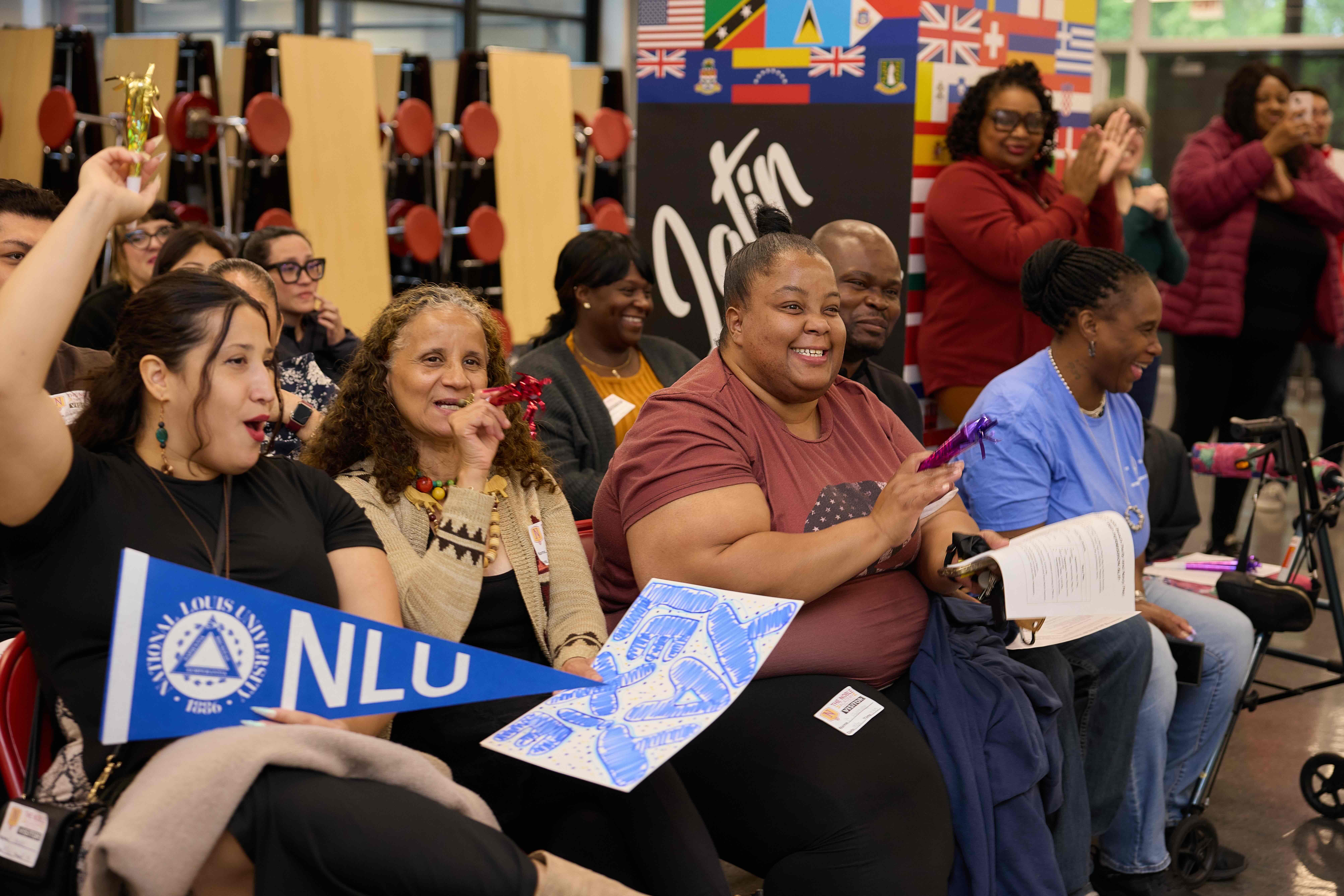 Parents and family members sit in the audience, cheering on their seniors. One holds an National Louis pennant. They all have party horns and some are blowing them.