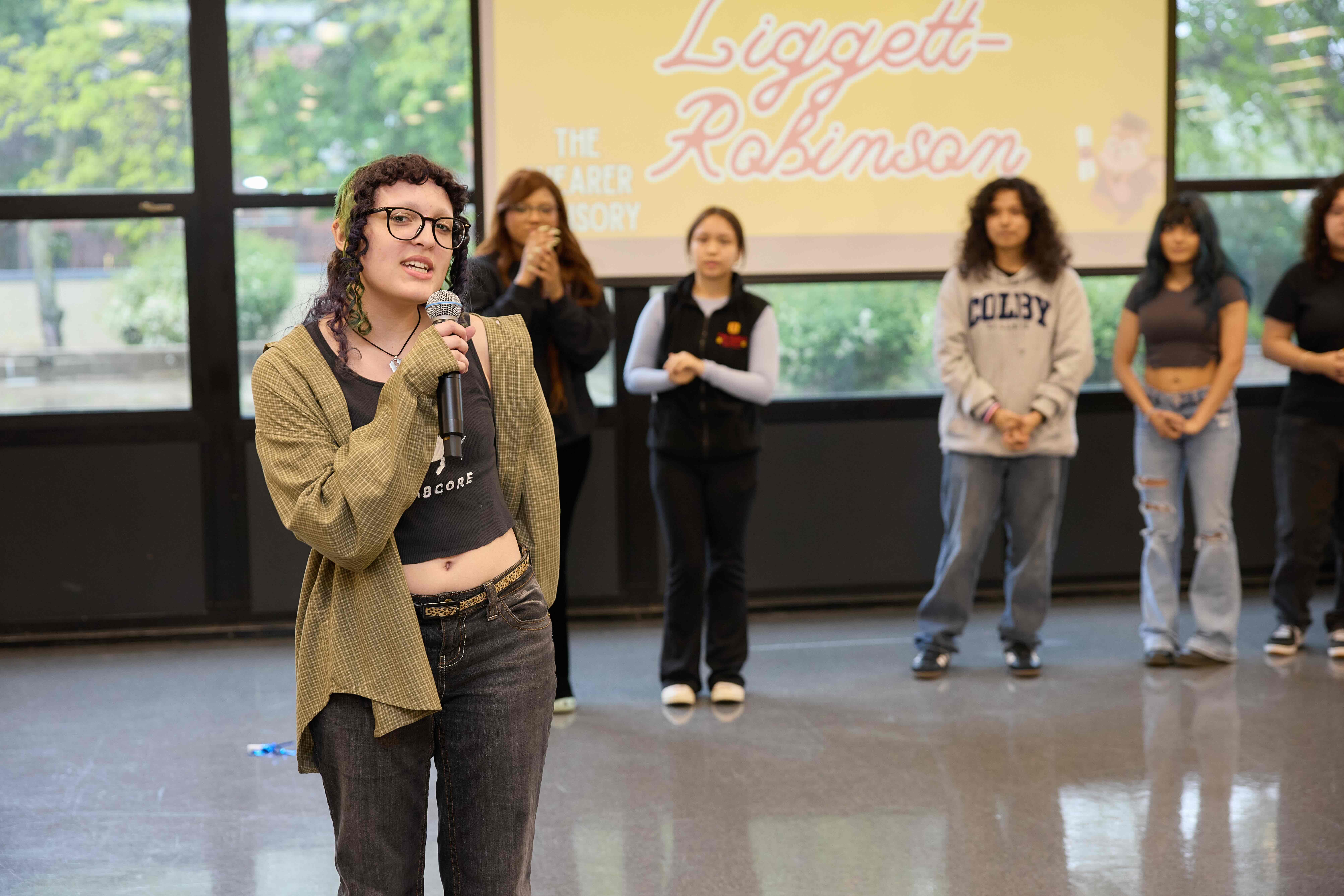 A TNA senior stands with a mic, announcing their college decision in front of a crowd of parents and peers.
