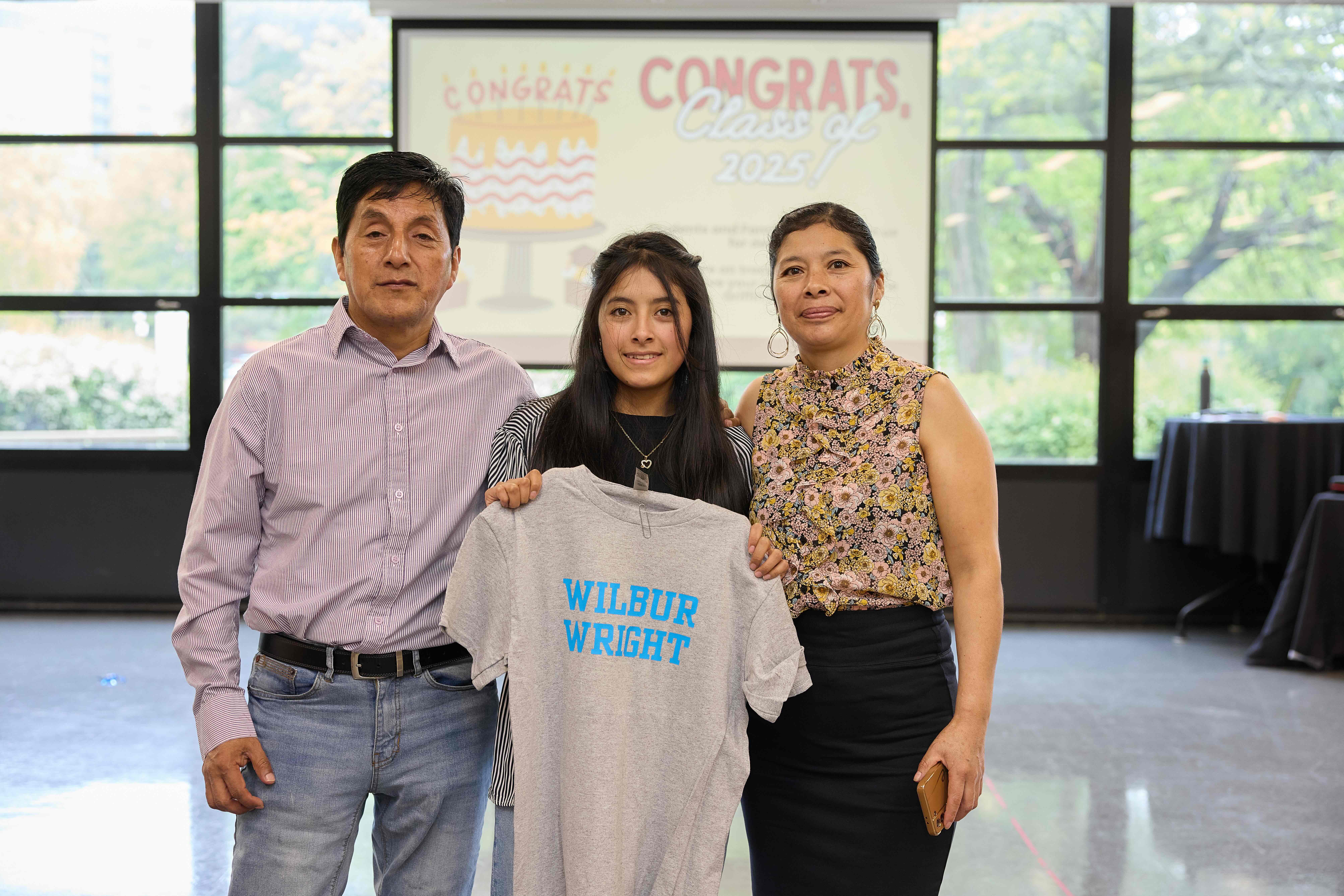 A TNA senior stands with their parents on both side. They are holding a gray t-shirt that says Wilbur Wright.