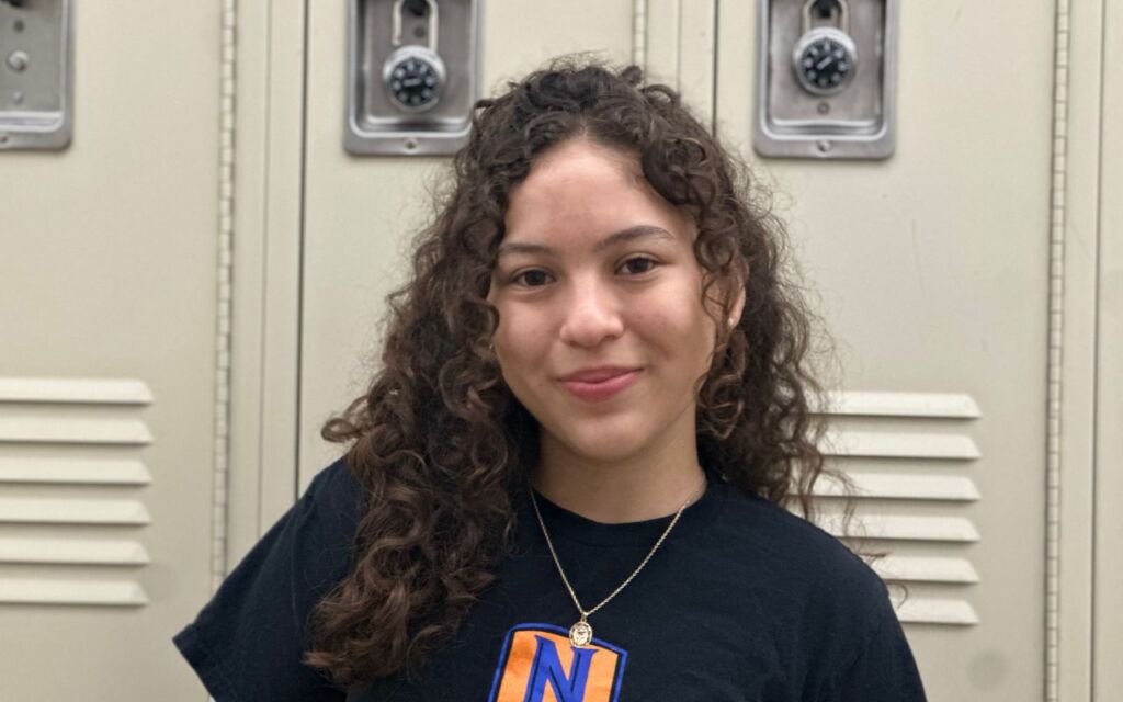 Ixchael Barajas, a student at Pritzker College Prep, smiles in front of school lockers. She is a young woman with long curly brown hair and light skin. She is wearing a black t-shirt with the Pritzker College Prep logo and a simple gold pendant necklace.
