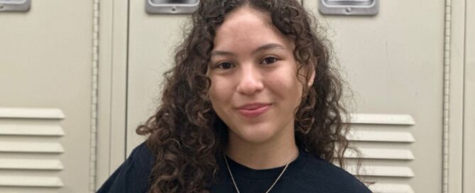 Ixchael Barajas, a student at Pritzker College Prep, smiles in front of school lockers. She is a young woman with long curly brown hair and light skin. She is wearing a black t-shirt with the Pritzker College Prep logo and a simple gold pendant necklace.