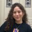 Ixchael Barajas, a student at Pritzker College Prep, smiles in front of school lockers. She is a young woman with long curly brown hair and light skin. She is wearing a black t-shirt with the Pritzker College Prep logo and a simple gold pendant necklace.