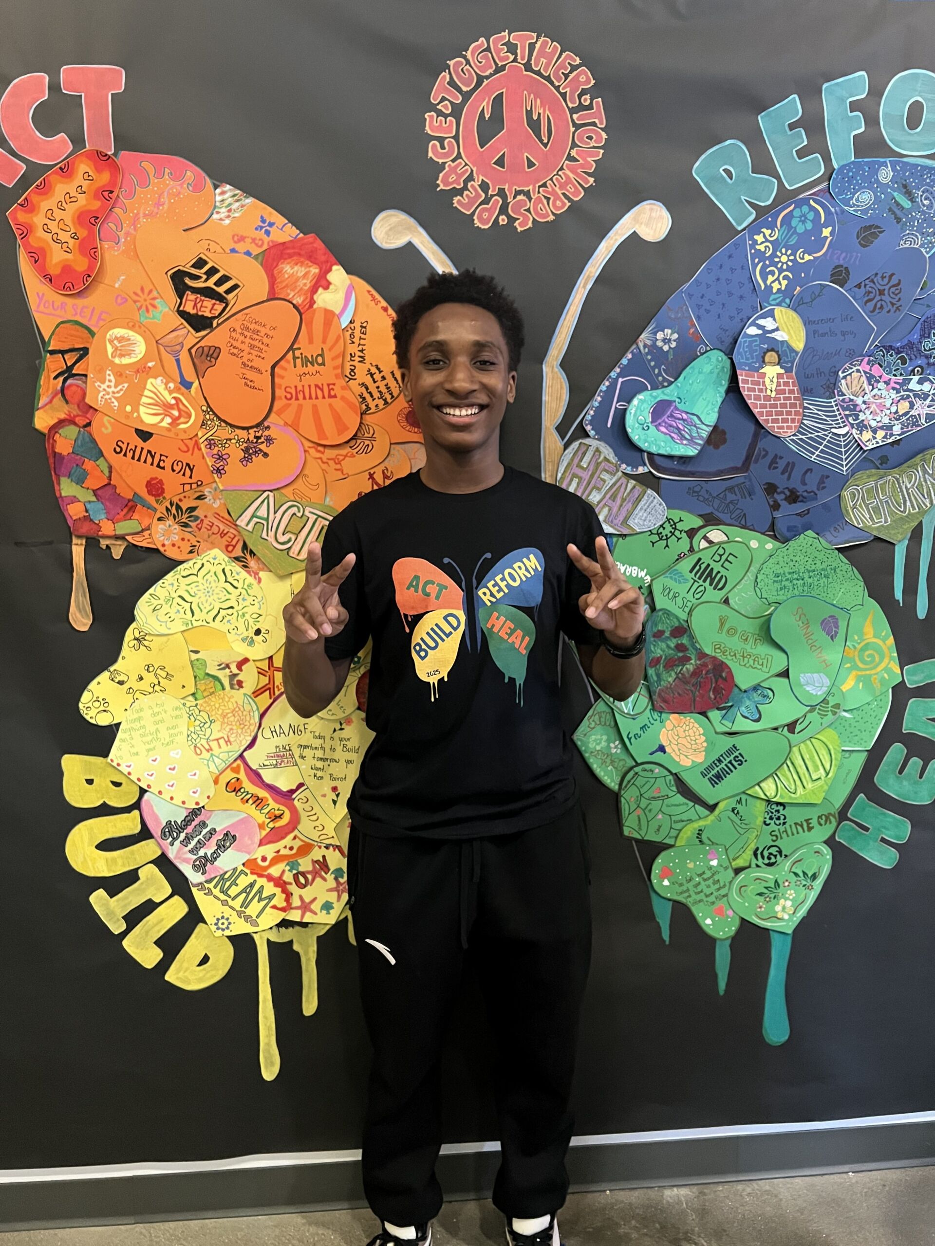 TTP Jamill butterfly photo Jamill throws up peace signs in front of a backdrop of a colorful butterfly. It looks like the wings are coming off of his back. He is a young man with short black curly hair and dark brown skin. He wears a black t-shirt with a matching butterfly on it. The butterfly is divided into four parts on each wing: the red upper left wing says "act", the yellow bottom left wing says "build", the blue upper right wing says "reform", and the green bottom right wing says "heal".