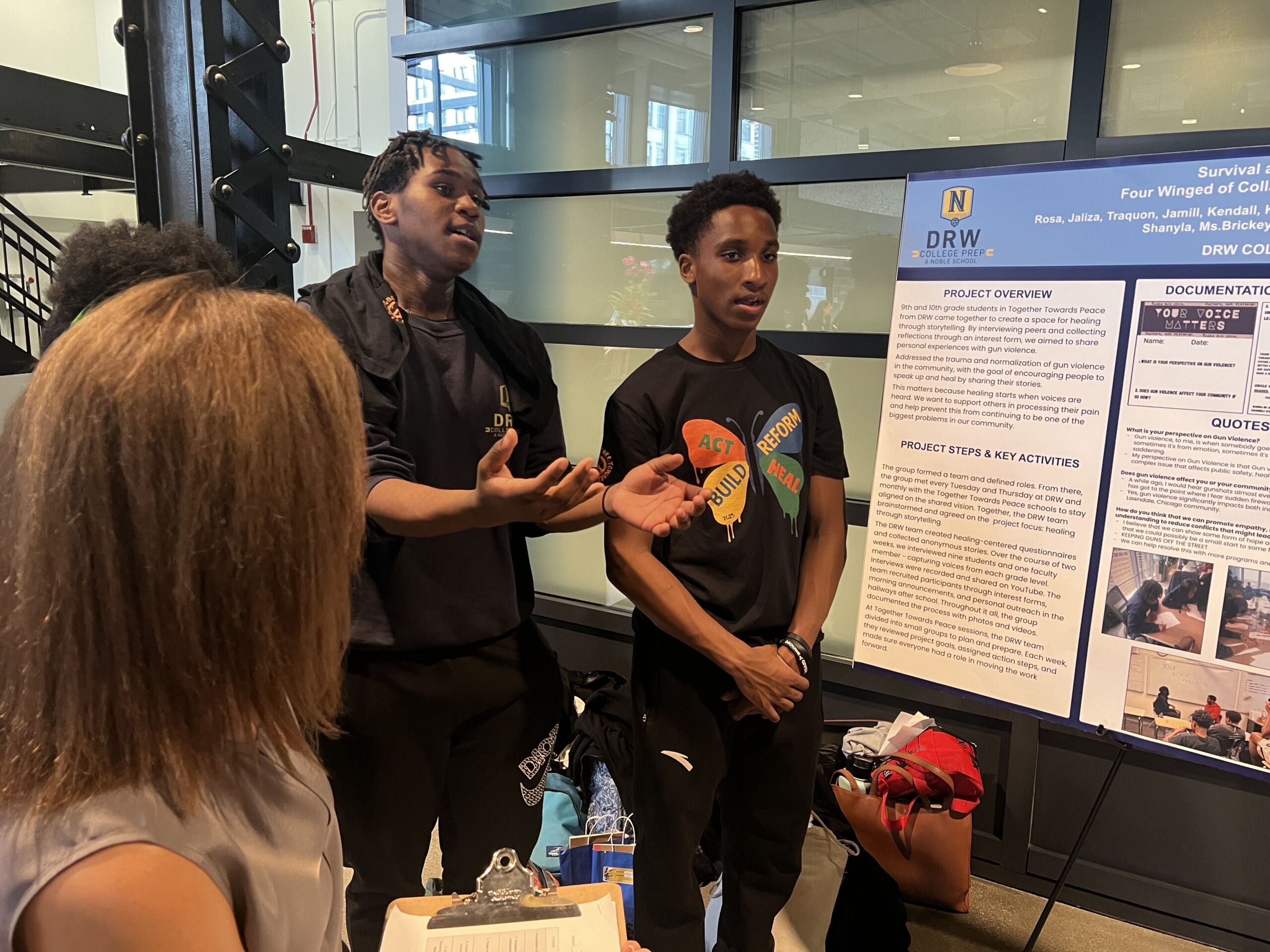 TTP Kendall presenting Kendall and Jamill stand to the side of a posterboard as they present their project. Kendall is talking with his hands emphasizing his point. He is a young man with short black locs and dark brown skin. He is wearing a black DRW t-shirt and jacket.