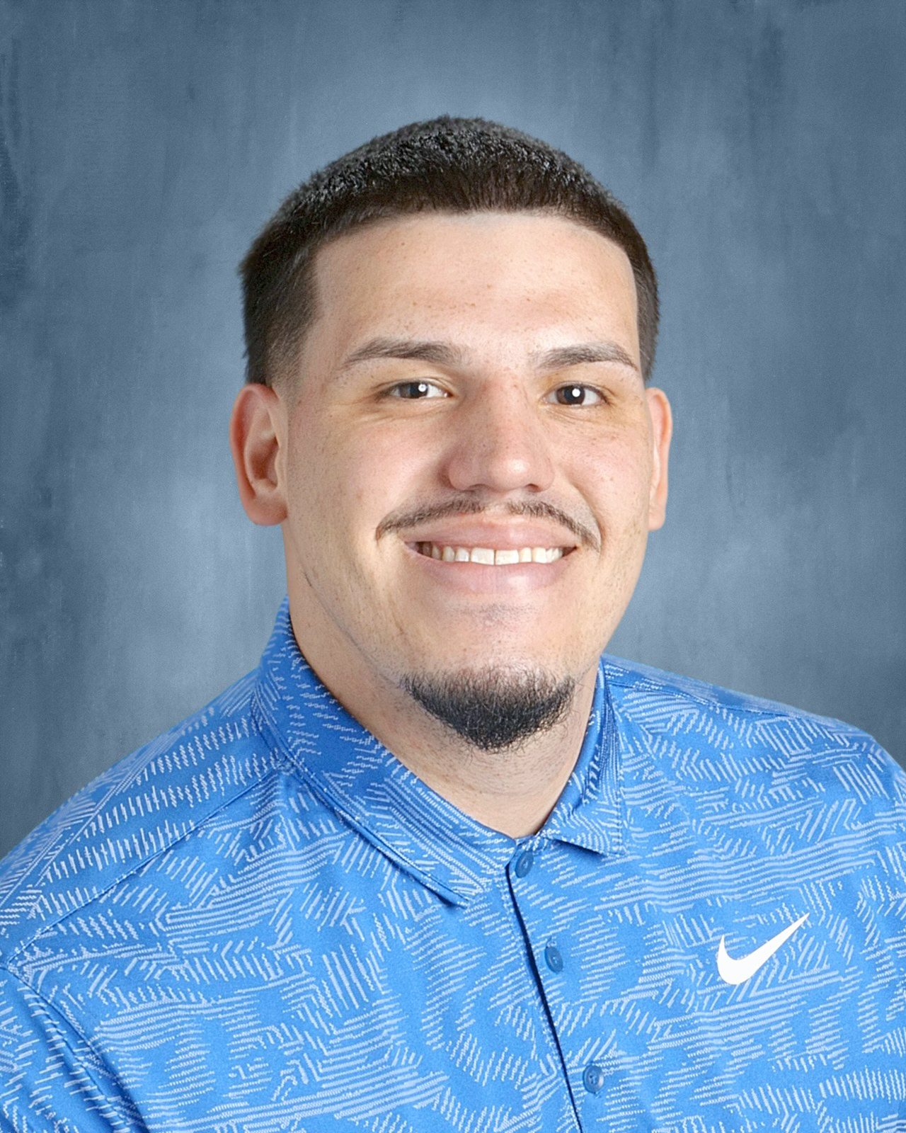 acosta A headshot of Coach Acosta. He is a man with short black buzzed hair, a goatee, and light skin. He is smiling and wearing a patterned light blue Nike button-up shirt. The background is a nondescript blue-gray pattern.