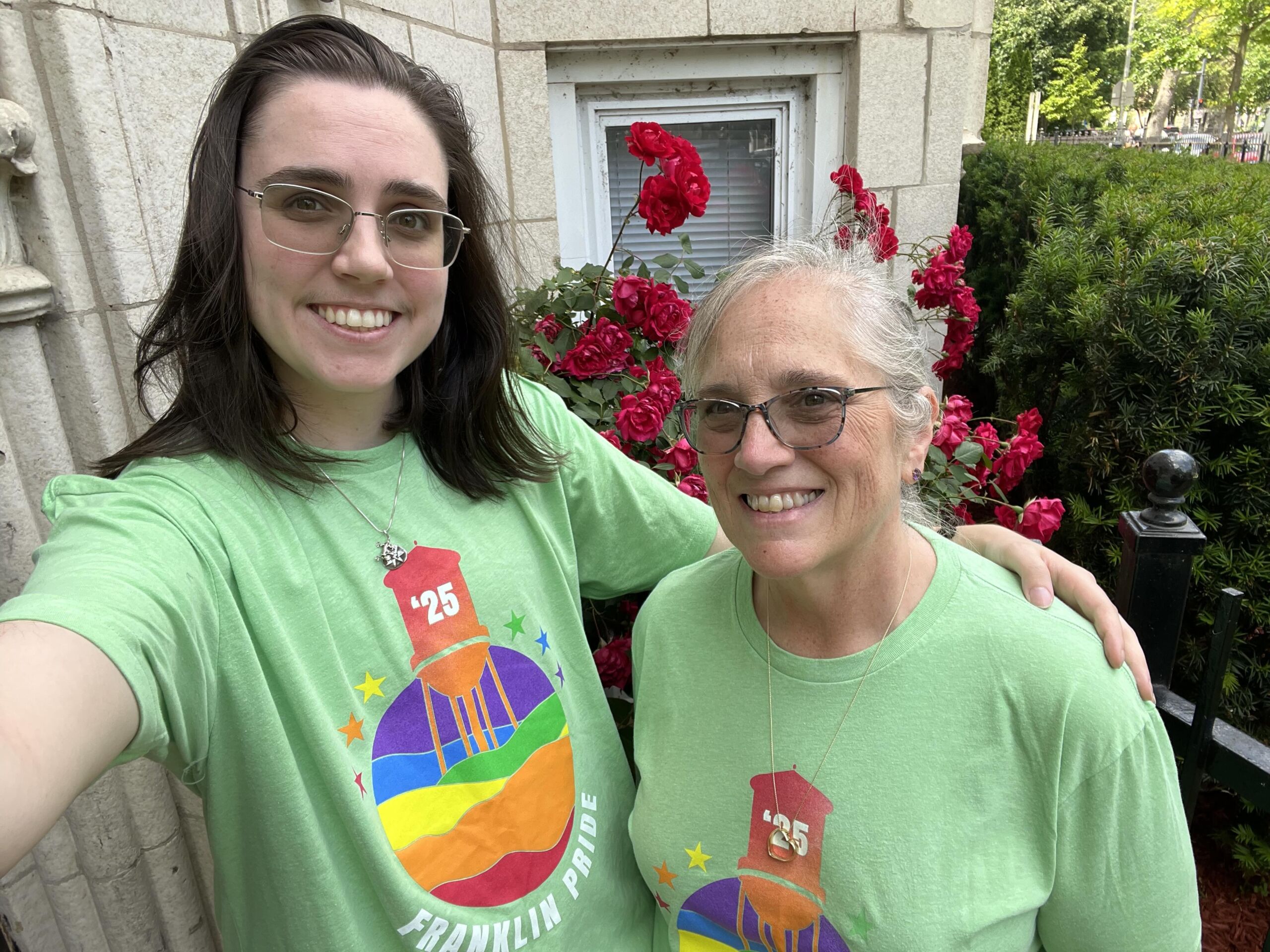 oliver pride blog 1 Mads Oliver stands next to their mom outside in front of a bush of roses. They are wearing matching light green Pride t-shirts and smiling. Mads is a white gender-nonconforming person with light skin, glasses, and medium length straight brown hair. Their mom is a white woman with light skin, glasses, and gray straight hair pulled back into a ponytail.
