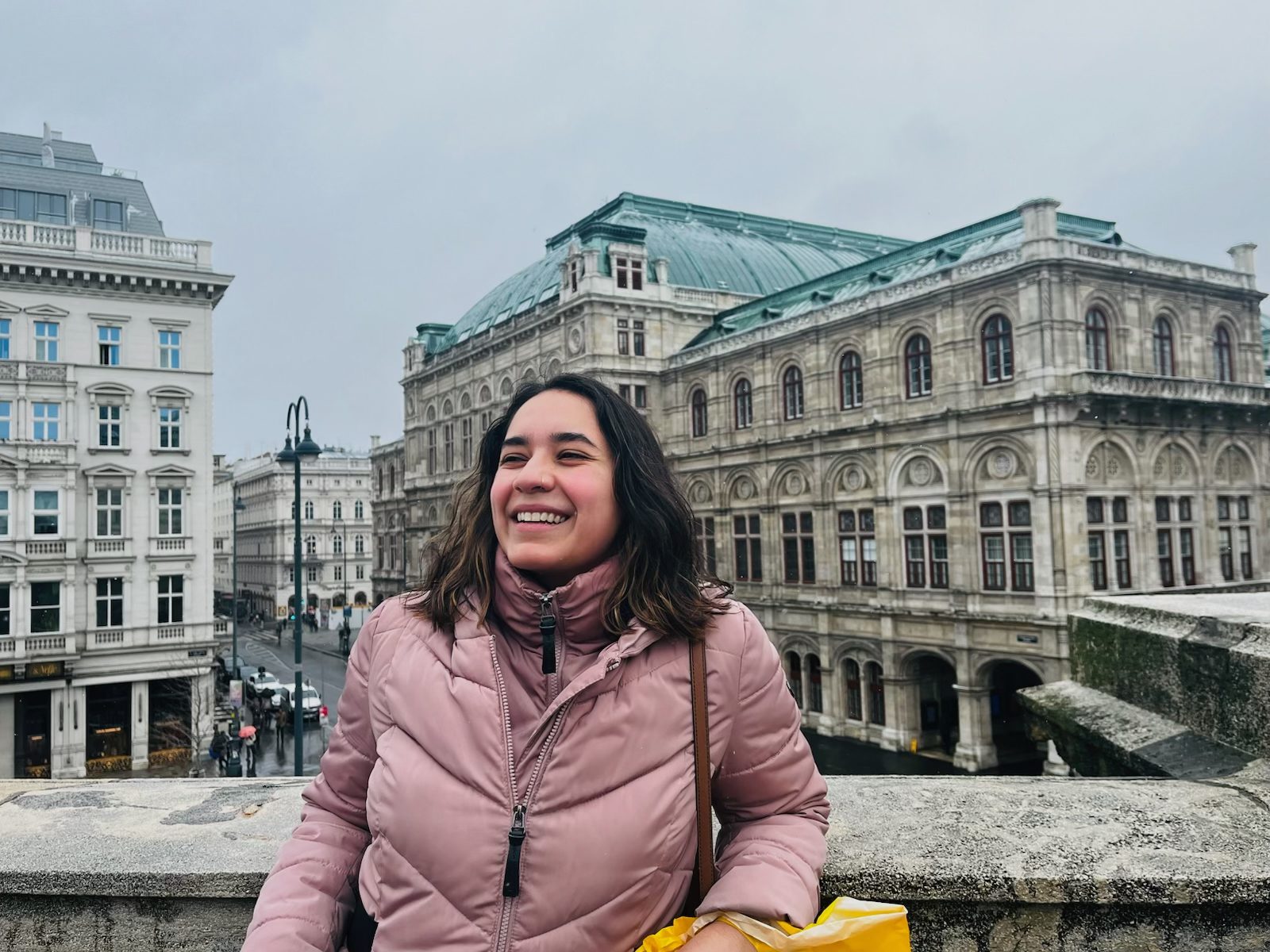 solis Coach Solis is smiling, standing on bridge in front a landscape of old, beautiful buildings. She is a woman with medium-length wavy brown hair and light skin. She is wearing a light pink puffer coat.