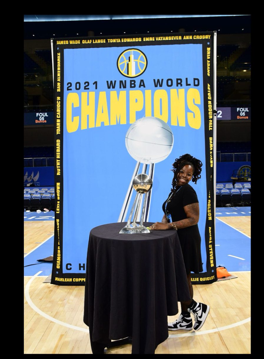 Jackson leans against a small covered cocktail table with the silver basketball WNBA championship trophy on it, in front of a large light blue photoshoot banner that has the yellow Chicago Sky logo on it and says "2021 WNBA World Champions". She is clearly on a basketball court and smiling brightly. She's dressed nicely in a simple short black professional dress and leggings. Her hair is up in a large ponytail of black locs.