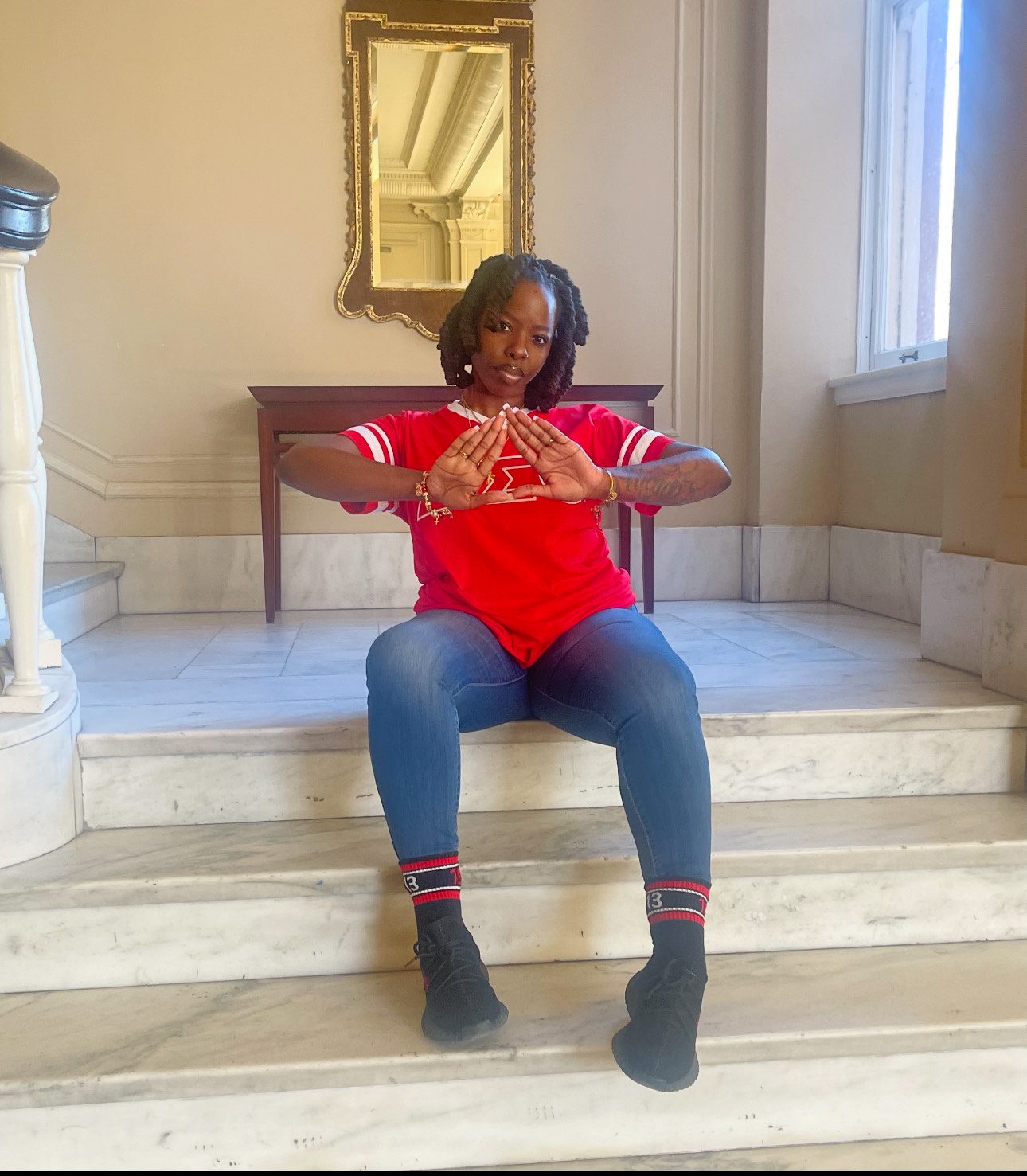 Jackson sits on marble stairs, putting her hands up to form a triangle between her fingers, the symbol of her sorority. She's serious and proud, looking confidently at the camera. Her giant black twists frame her face, ending right at her chin. She is wearing a bright red jersey with her sorority's Greek letters on it, blue jeans, matching red and black socks, and black tennis shoes.