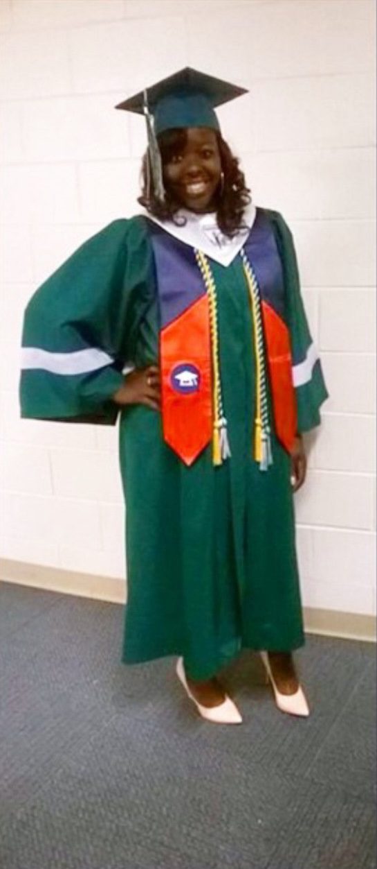Jackson at her Gary Comer College Prep graduation. She is posing for a photo in her dark green Comer robes and graduation cap. She has two tassels and a bright orange and blue sash. Loose dark brown curls that reach just past her chin frame her smiling face.