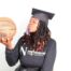 Autumn Jackson, a Gary Comer College Prep alum, holds a basketball and blows a kiss towards it. She is a young Black woman with medium-light brown skin and black locs that flow down past her shoulders and end in a bright burnt orange color. She is wearing a black graduation cap and a black sweatshirt that says "Northwestern Sports Administration" and has the big N Northwestern University logo on it.