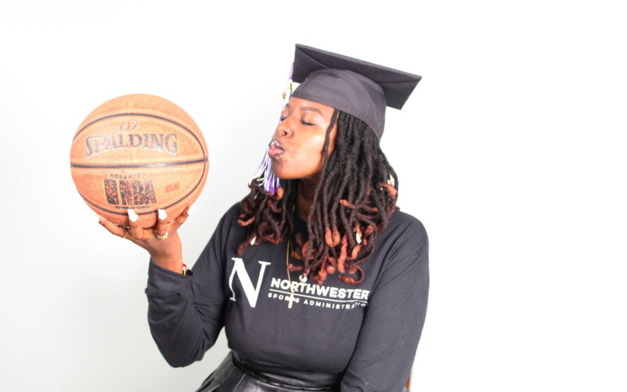 Autumn Jackson, a Gary Comer College Prep alum, holds a basketball and blows a kiss towards it. She is a young Black woman with medium-light brown skin and black locs that flow down past her shoulders and end in a bright burnt orange color. She is wearing a black graduation cap and a black sweatshirt that says "Northwestern Sports Administration" and has the big N Northwestern University logo on it.