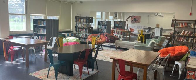 Johnson College Prep's in-house library is full of tables, plush chairs, bean bags, and bookcases. It is well lit and looks cozy with rugs spread out all across the room. A large mirror is on the wall, reflecting most of the spacious library.