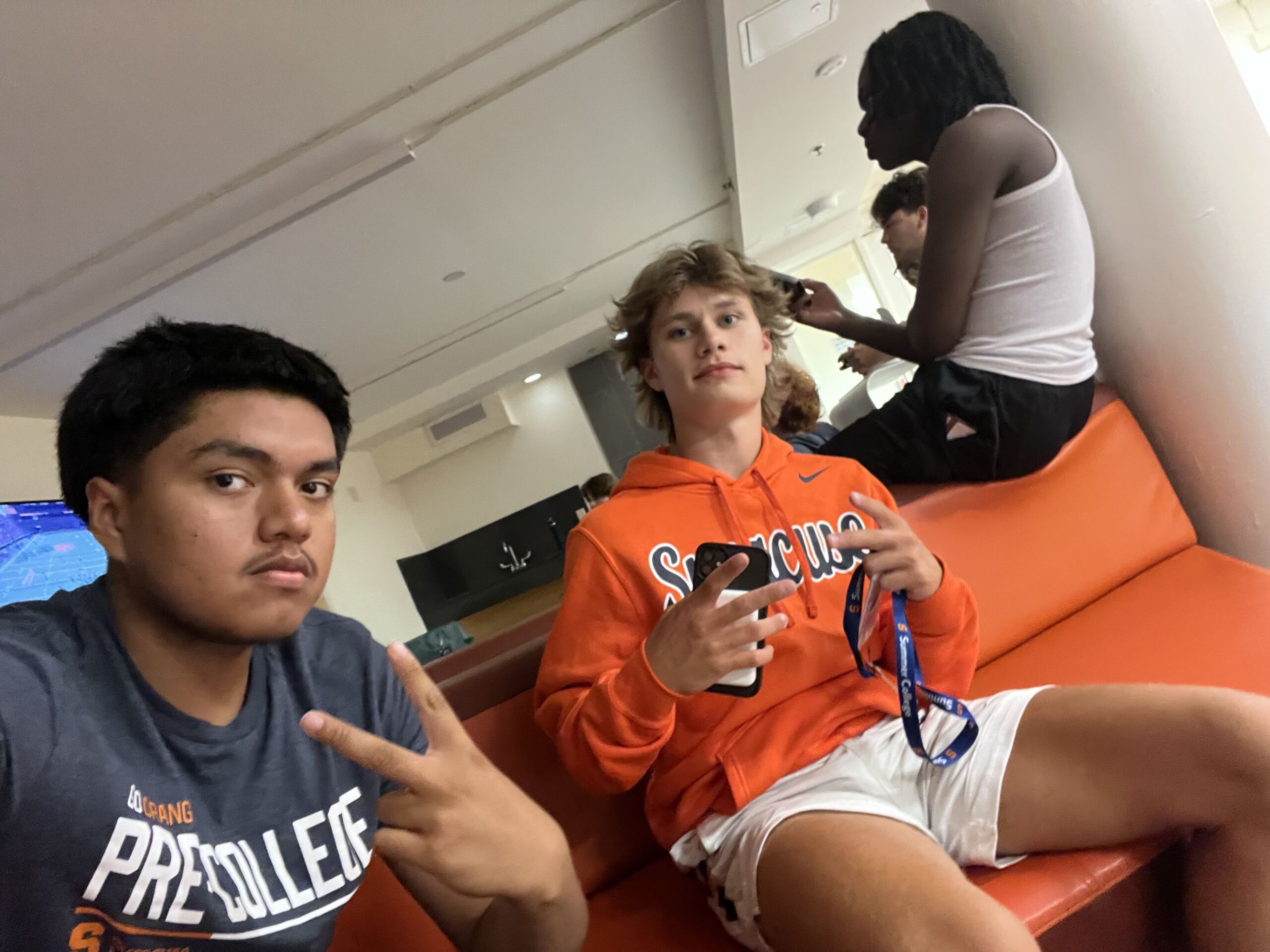 Juan Gonzalez Speer SOL Juan sits on a bright orange sofa in a Syracruse University dorm common space with other students. He is a young man with short black hair and brown skin. He is wearing a gray t-shirt that says "Pre-College" on it and he is throwing up a peace sign.
