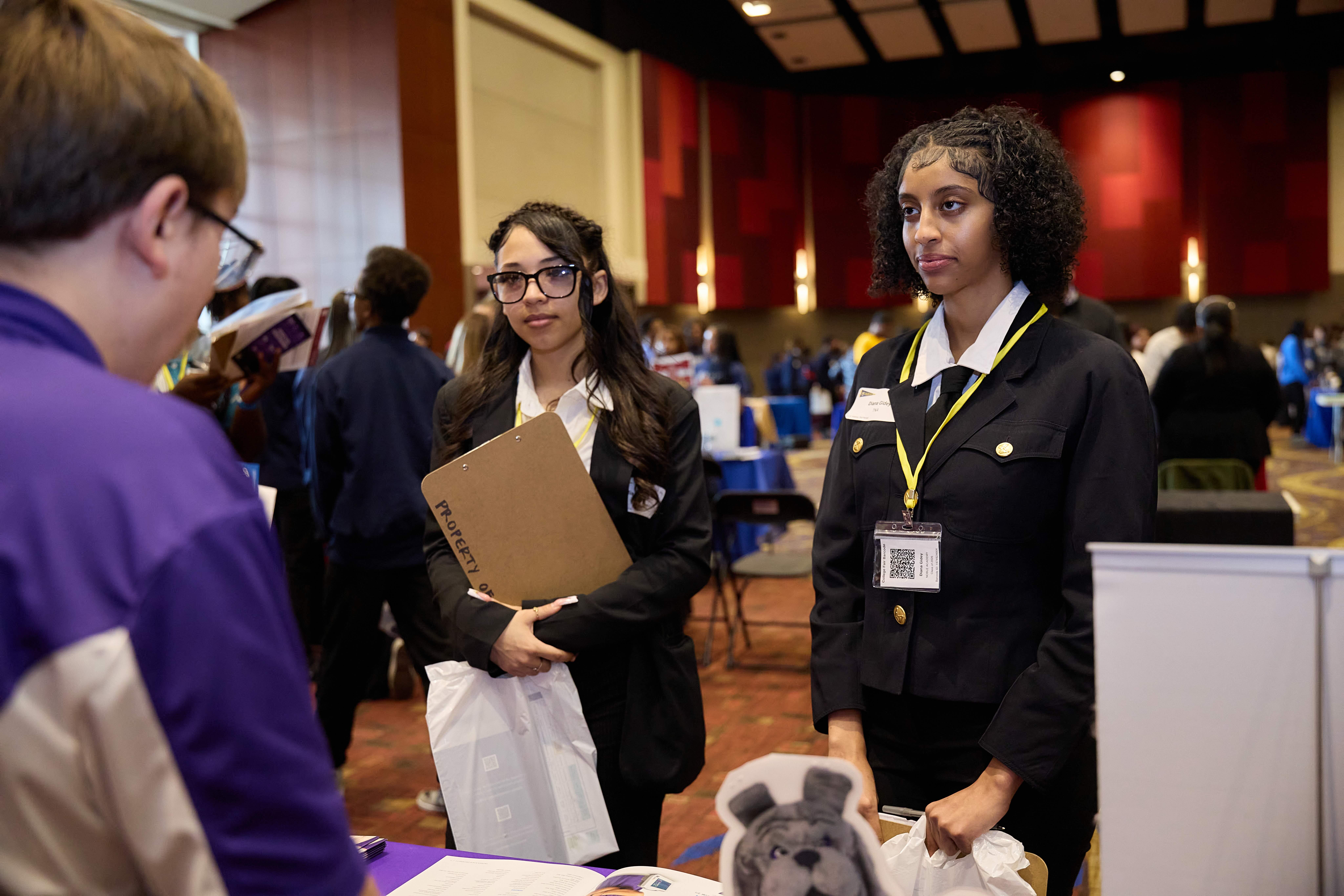 tna students sy25 college fair Two TNA students stand in front of a college representative at the Noble Junior College Fair. They are both young woman dressed up in smart black dress suits. They are paying close attention to the college representative. The student on the right is a young woman with dark brown skin and brown hair that is braided on the top and then let out into a short natural tight curl style. The student on the left is a young woman with light brown skin and long brown straight hair with a single braid on top and glasses