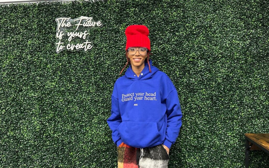 Chelsea Billingsley, crochet artist and Chicago Bulls College Prep alum, stands in front of a leafy backdrop with her hands in her pockets, smiling. She is a Black woman with medium length red locs and brown skin. she is wearing a bright blue sweatshirt that says "Protect your head. Guard your heart." and a bright red tall beanie that she crocheted. To the left of her head, you can see a fluorescent sign that says "The Future is yours to create".