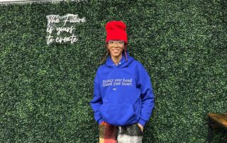 Chelsea Billingsley, crochet artist and Chicago Bulls College Prep alum, stands in front of a leafy backdrop with her hands in her pockets, smiling. She is a Black woman with medium length red locs and brown skin. she is wearing a bright blue sweatshirt that says "Protect your head. Guard your heart." and a bright red tall beanie that she crocheted. To the left of her head, you can see a fluorescent sign that says "The Future is yours to create".