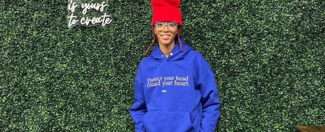 Chelsea Billingsley, crochet artist and Chicago Bulls College Prep alum, stands in front of a leafy backdrop with her hands in her pockets, smiling. She is a Black woman with medium length red locs and brown skin. she is wearing a bright blue sweatshirt that says "Protect your head. Guard your heart." and a bright red tall beanie that she crocheted. To the left of her head, you can see a fluorescent sign that says "The Future is yours to create".