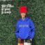 Chelsea Billingsley, crochet artist and Chicago Bulls College Prep alum, stands in front of a leafy backdrop with her hands in her pockets, smiling. She is a Black woman with medium length red locs and brown skin. she is wearing a bright blue sweatshirt that says "Protect your head. Guard your heart." and a bright red tall beanie that she crocheted. To the left of her head, you can see a fluorescent sign that says "The Future is yours to create".