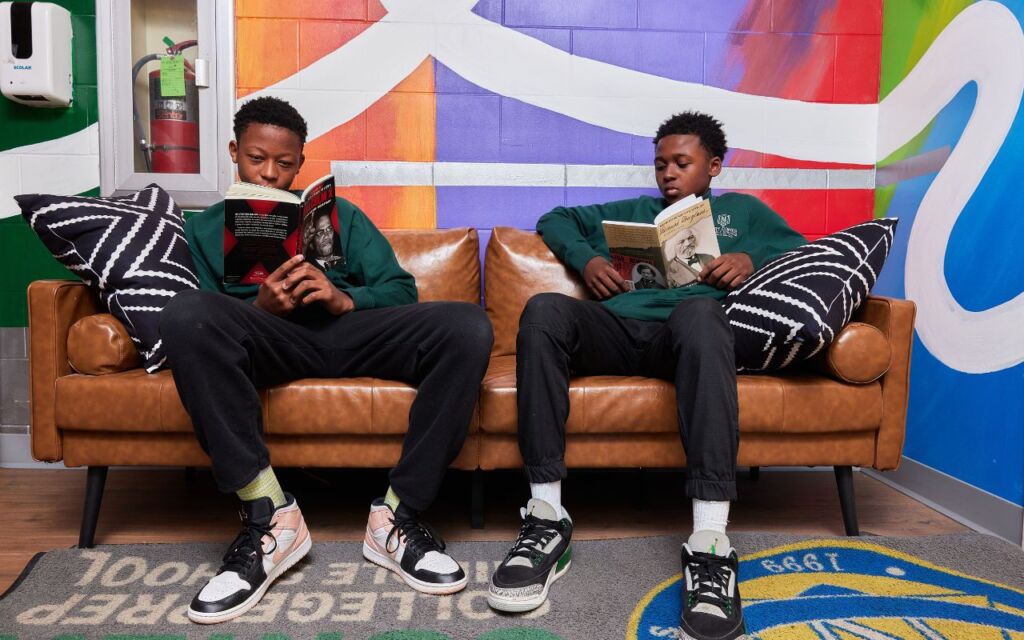 Two students sit on a leather couch in front of a colorful mural. They are both reading two different books: one about Malcolm X and the other about Frederick Douglass.
