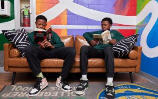Two students sit on a leather couch in front of a colorful mural. They are both reading two different books: one about Malcolm X and the other about Frederick Douglass.