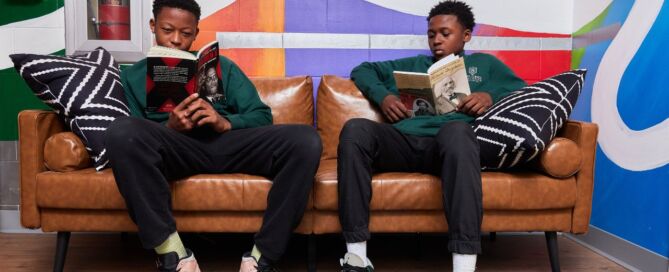 Two students sit on a leather couch in front of a colorful mural. They are both reading two different books: one about Malcolm X and the other about Frederick Douglass.