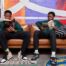 Two students sit on a leather couch in front of a colorful mural. They are both reading two different books: one about Malcolm X and the other about Frederick Douglass.