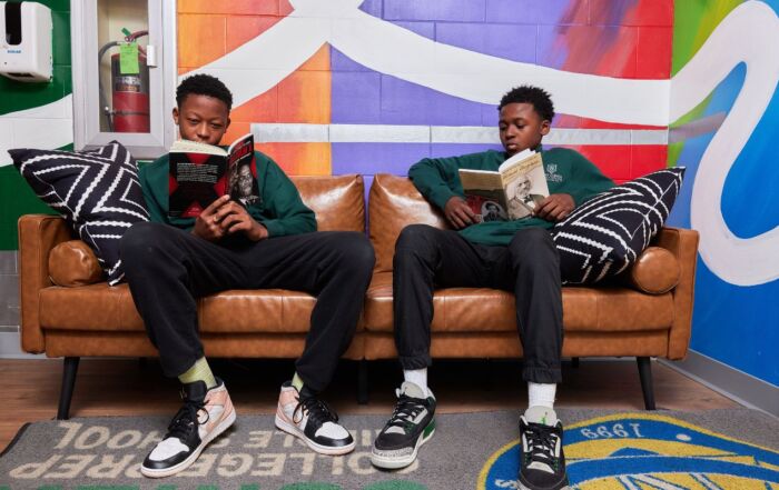 Two students sit on a leather couch in front of a colorful mural. They are both reading two different books: one about Malcolm X and the other about Frederick Douglass.
