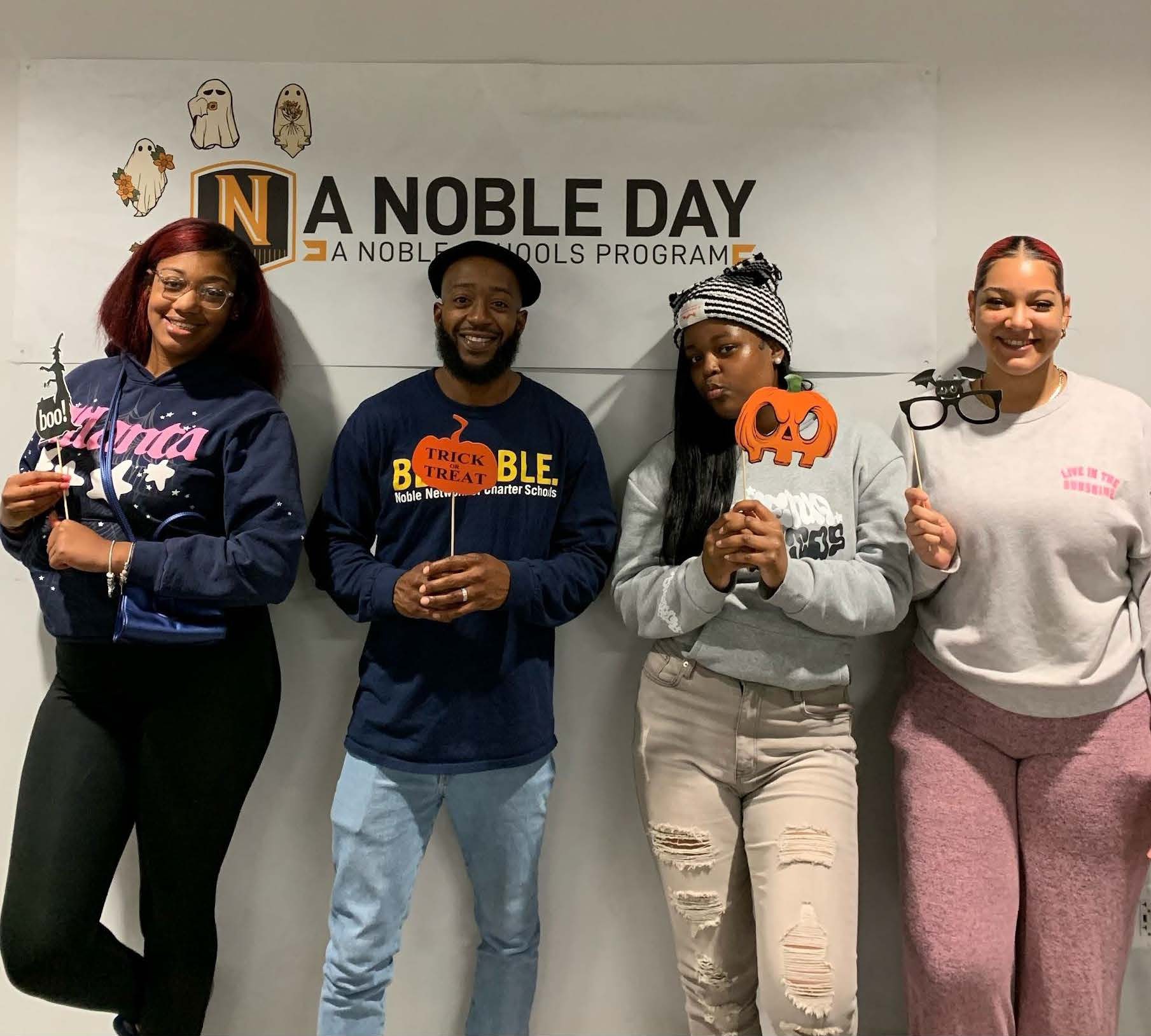 Justin Harris stands with three Baker alumni in front of a sign that says "A Noble Day, A Noble Schools Program". They are smiling and holding little Halloween-themed mini signs. Harris is a Black man who is bald with a medium-length black beard and medium brown skin. He is wearing a flat cap and a dark blue "Be Noble" sweatshirt.