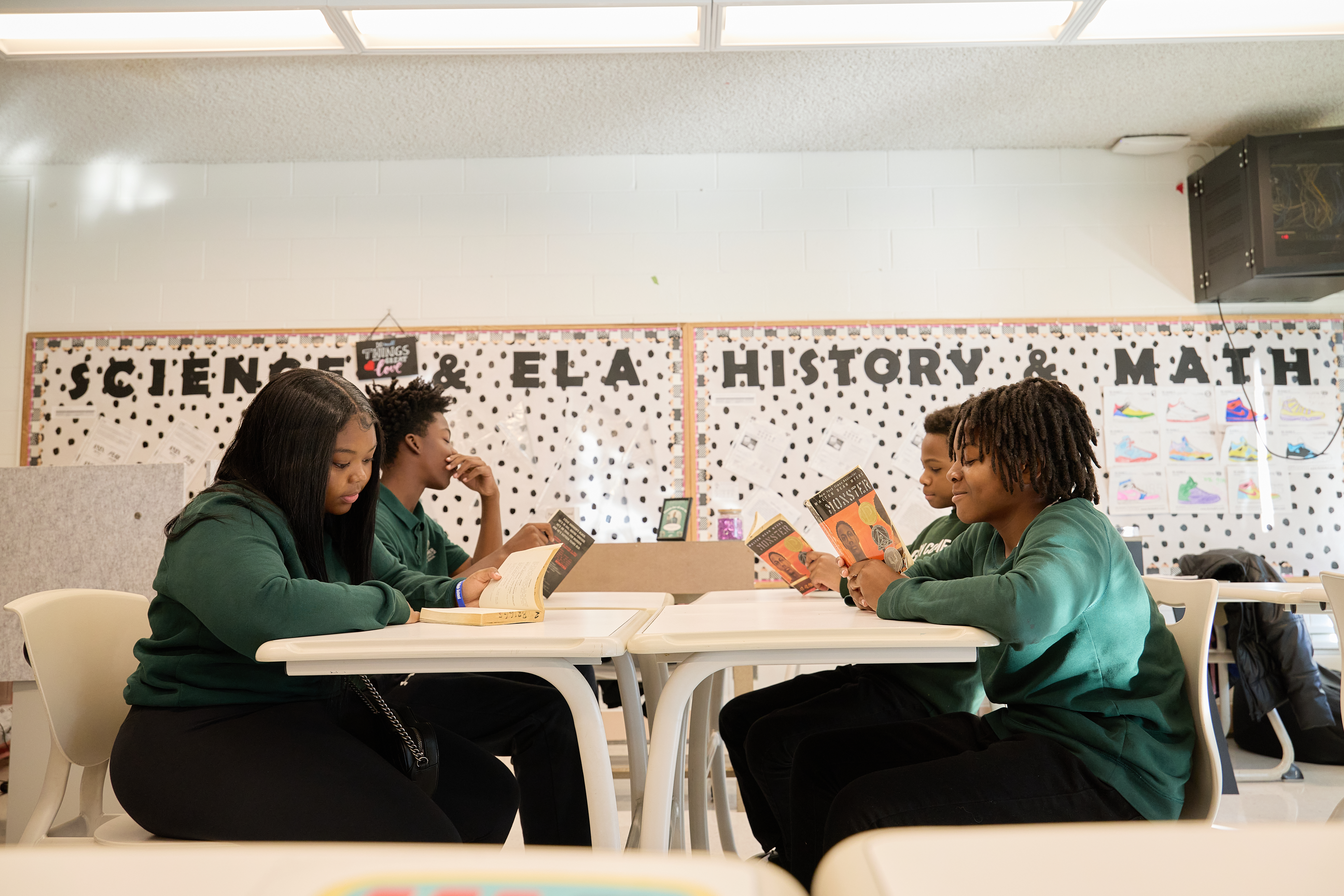 _LM_2049 Four students sit at a classroom desks pushed together into a table. All of them are reading the same book.